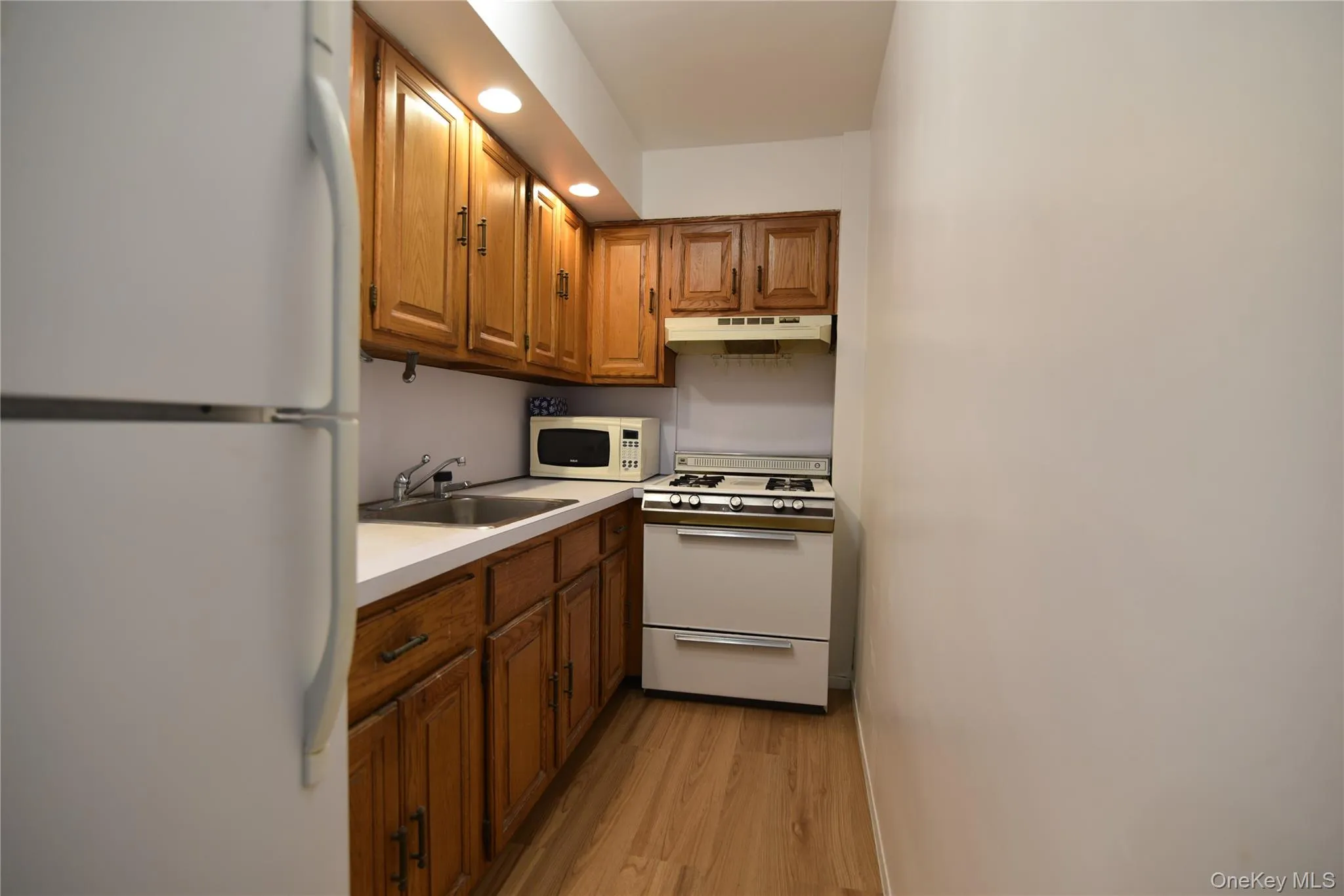 Kitchen with white appliances, brown cabinetry, light countertops, under cabinet range hood, and light wood-type flooring Kitchen with white appliances, brown cabinetry, light countertops, under cabinet range hood, and light wood-type flooring