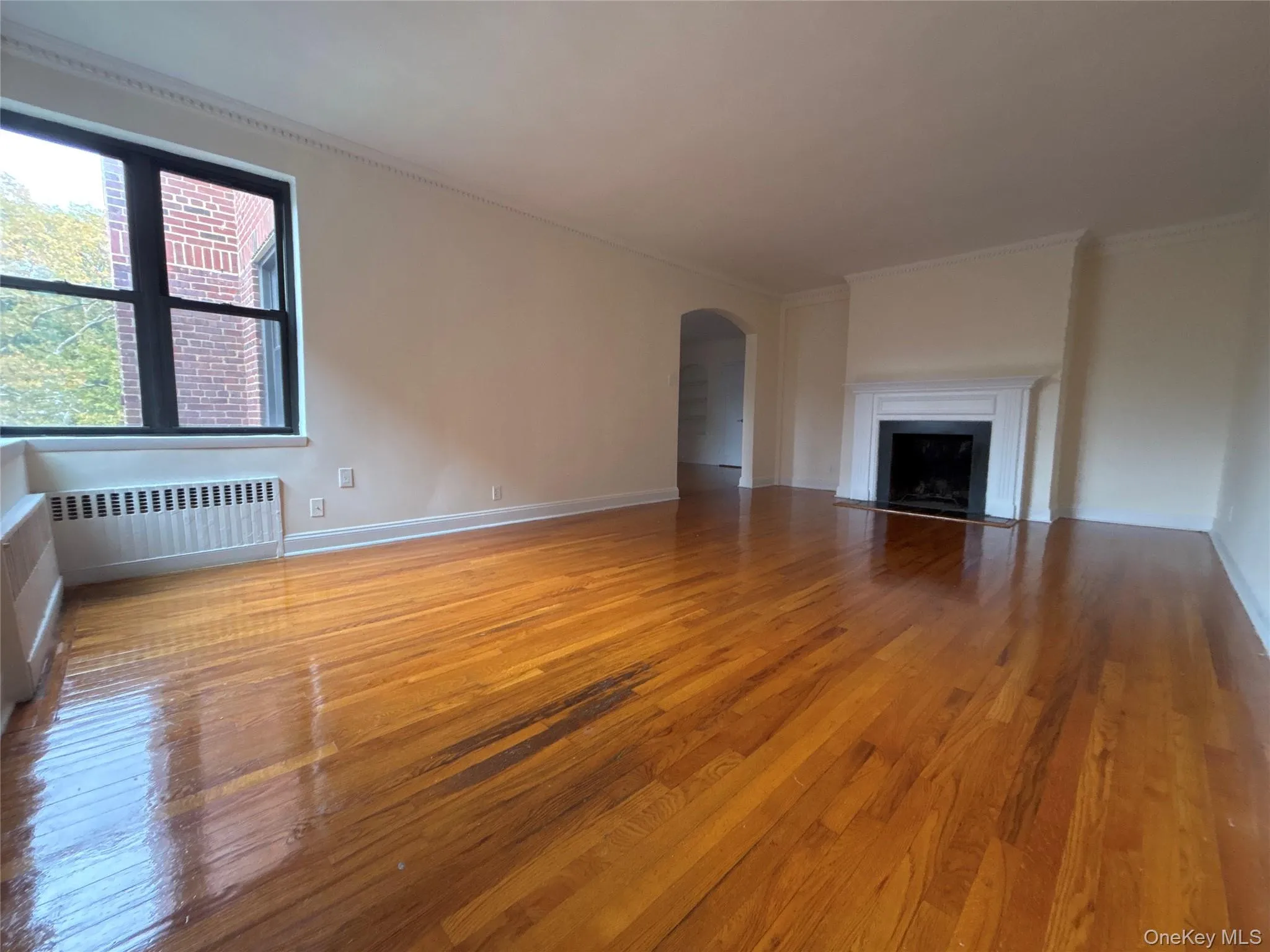 Unfurnished living room with polished hardwood flooring, radiator, arched walkways, a fireplace with flush hearth, and ornamental molding Unfurnished living room with polished hardwood flooring, radiator, arched walkways, a fireplace with flush hearth, and ornamental molding