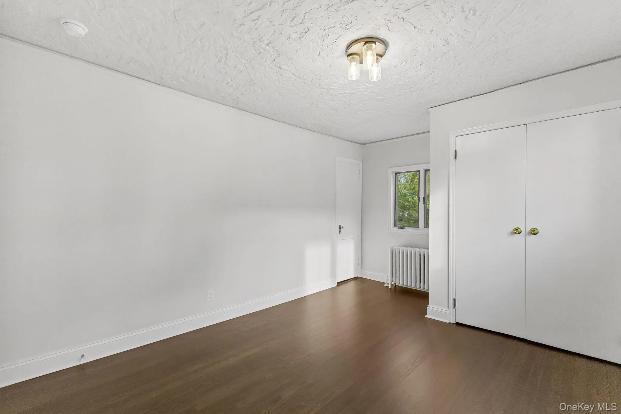 Unfurnished bedroom featuring radiator heating unit, dark wood-style floors, a closet, and a textured ceiling Unfurnished bedroom featuring radiator heating unit, dark wood-style floors, a closet, and a textured ceiling