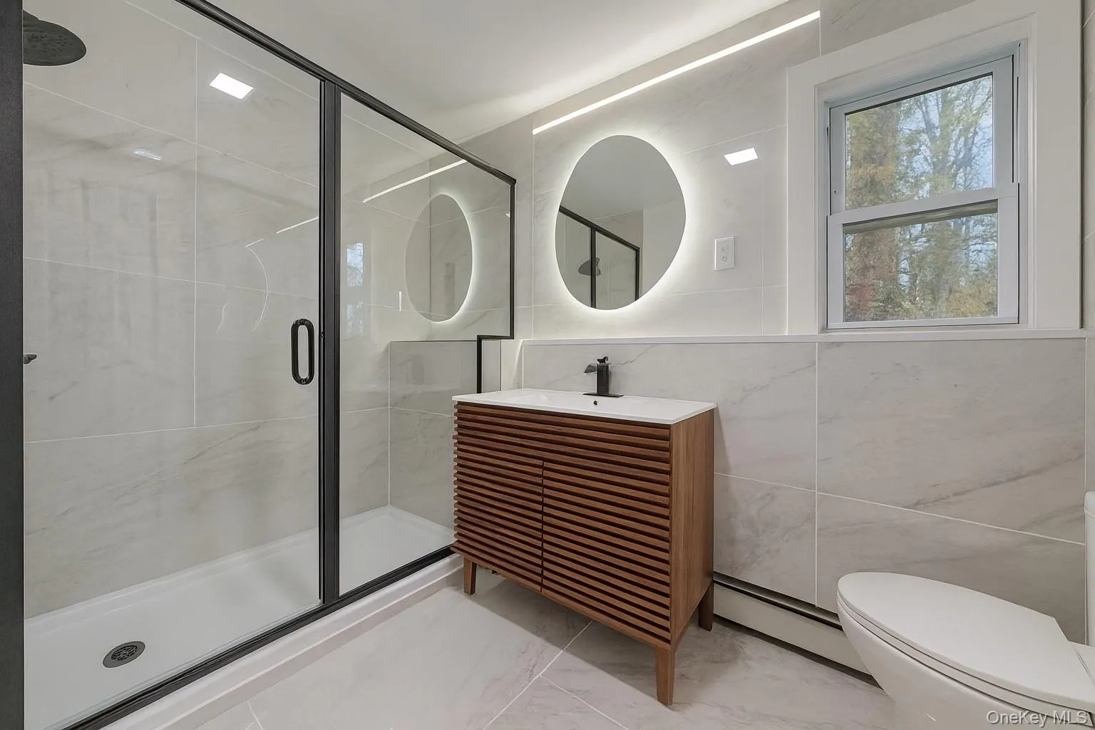 Main level full bath with vanity, a shower stall, tile walls, baseboard heating, and light marble finish flooring Main level full bath with vanity, a shower stall, tile walls, baseboard heating, and light marble finish flooring