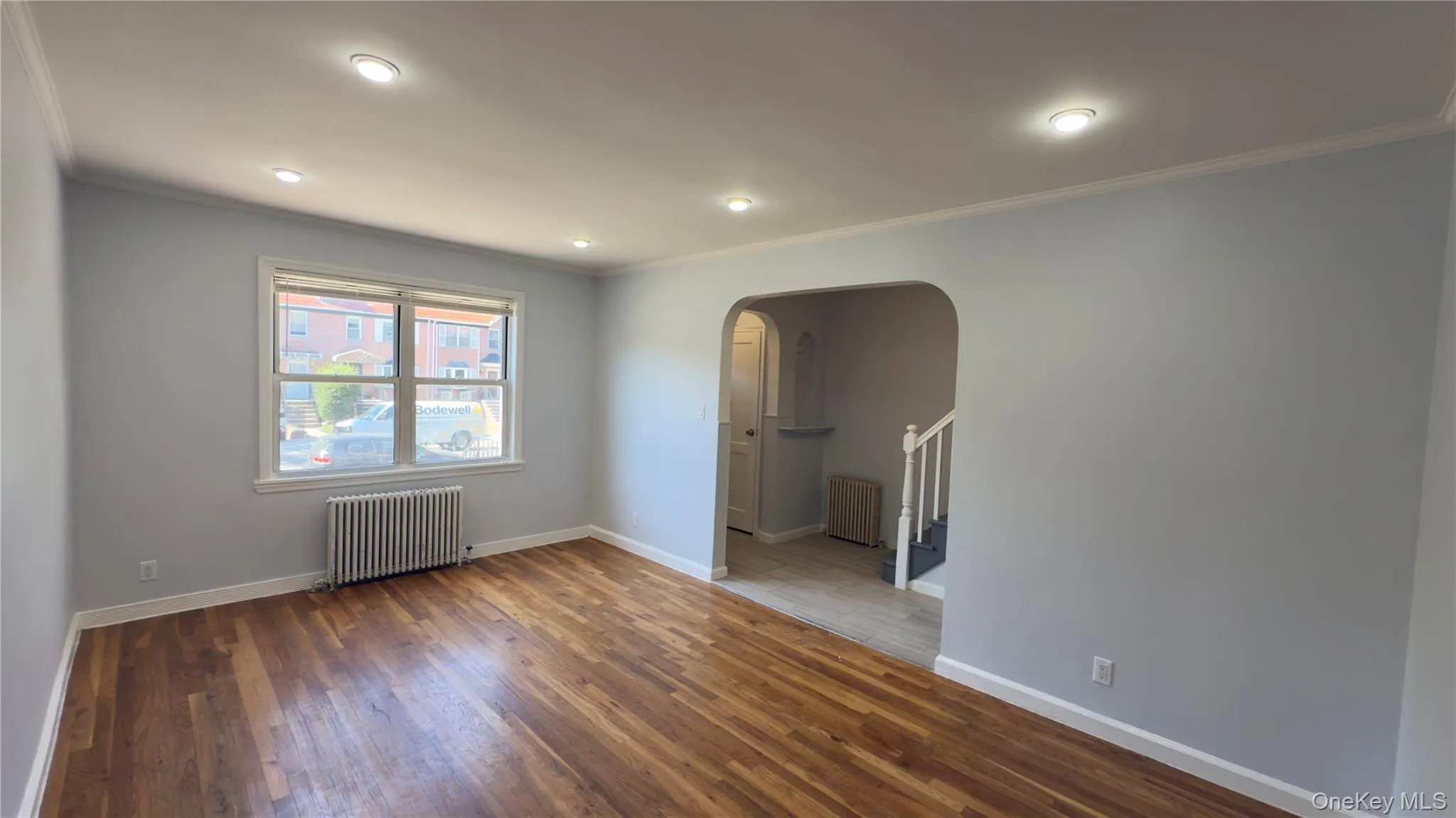 Unfurnished room featuring arched walkways, ornamental molding, radiator heating unit, wood finished floors, and stairs Unfurnished room featuring arched walkways, ornamental molding, radiator heating unit, wood finished floors, and stairs