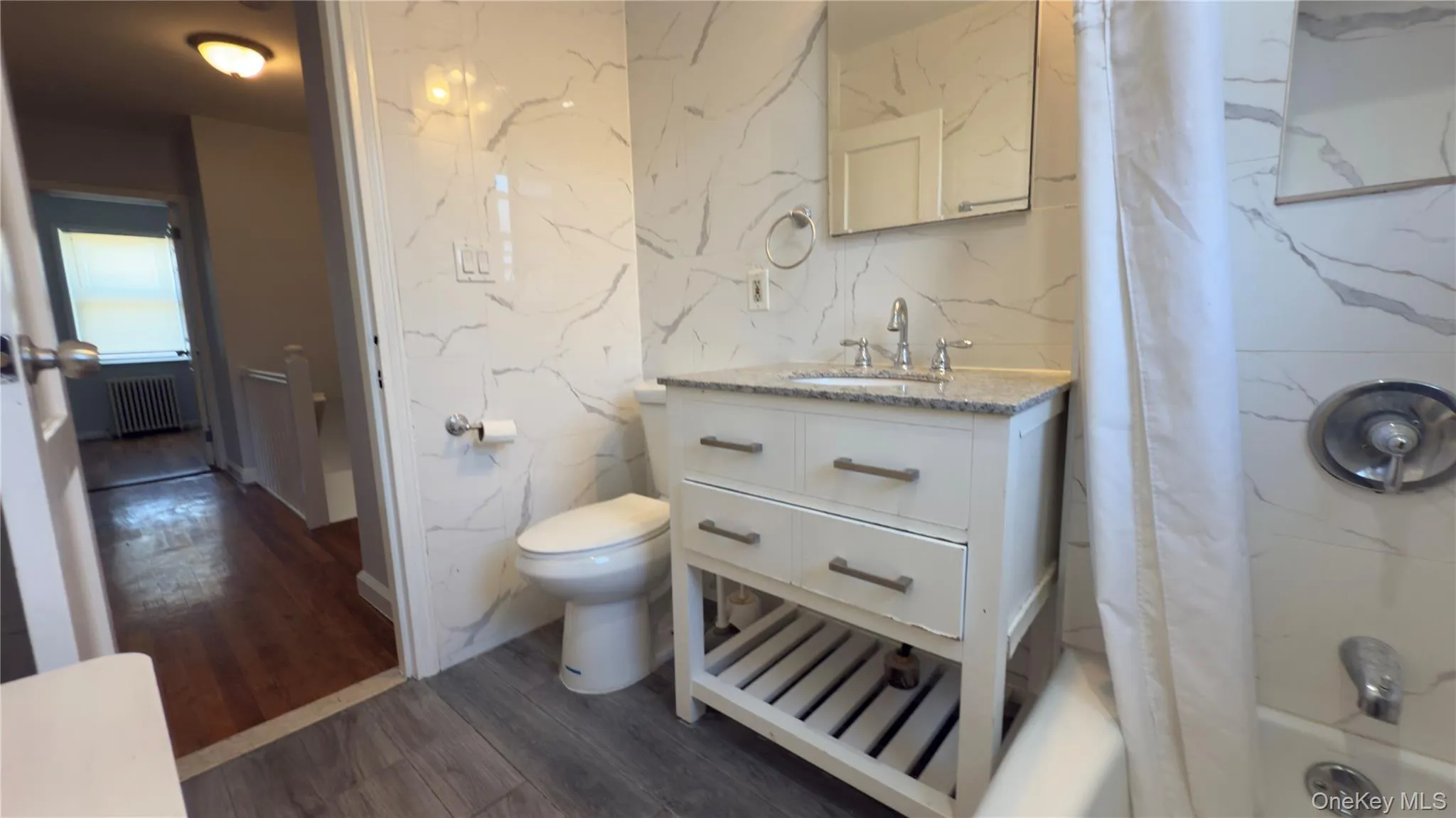 Full bath featuring shower / bath combination with curtain, vanity, dark wood-style floors, radiator heating unit, and tile walls Full bath featuring shower / bath combination with curtain, vanity, dark wood-style floors, radiator heating unit, and tile walls