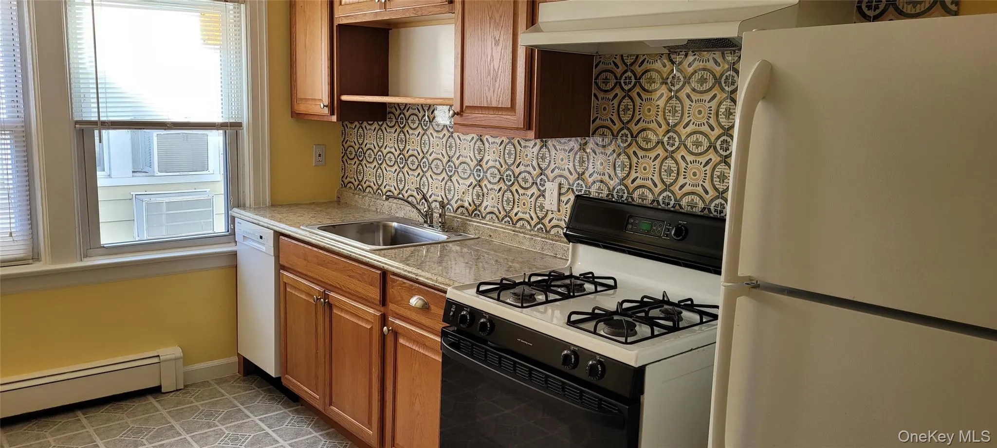 Kitchen with white appliances, decorative backsplash, a baseboard radiator, light countertops, and brown cabinetry Kitchen with white appliances, decorative backsplash, a baseboard radiator, light countertops, and brown cabinetry