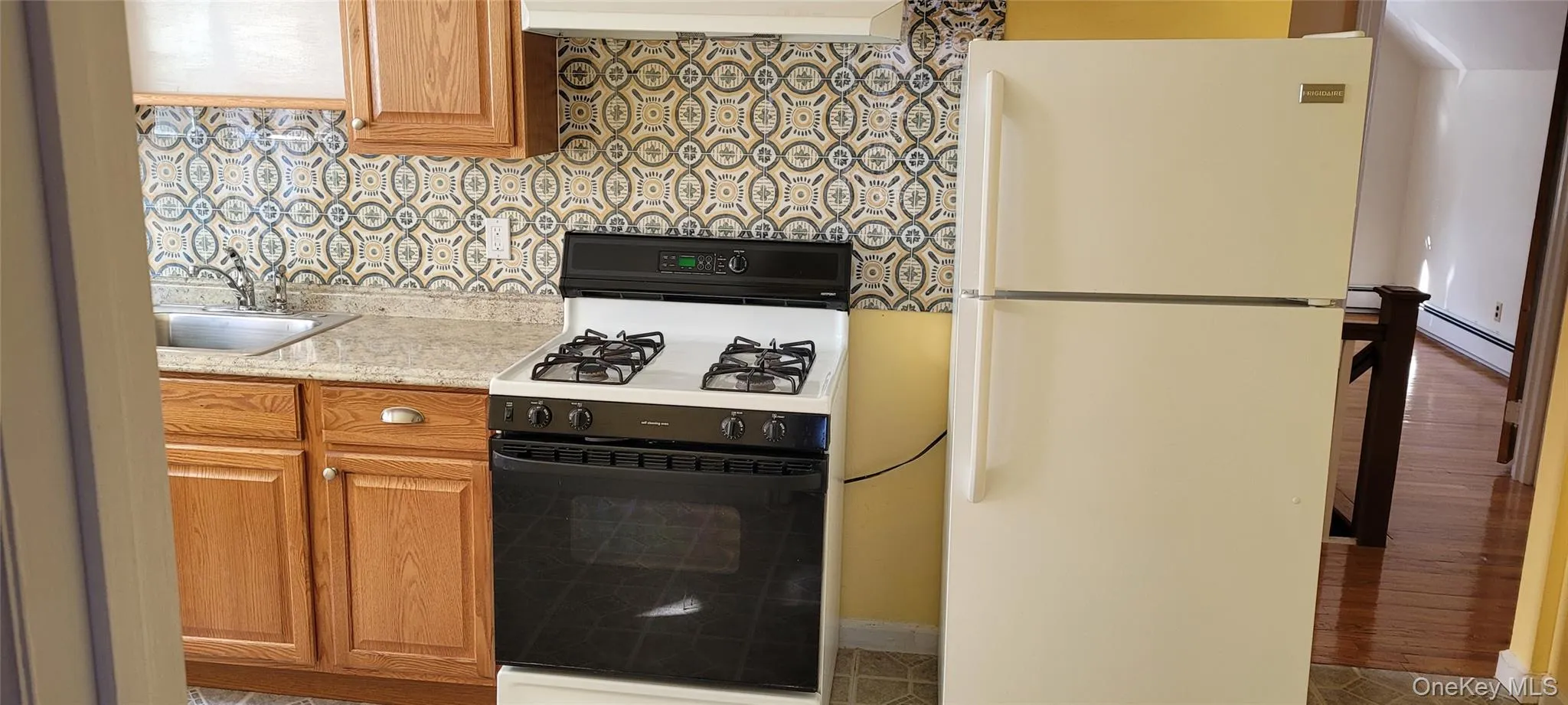 Kitchen featuring gas range oven, freestanding refrigerator, brown cabinetry, backsplash, and a baseboard heating unit Kitchen featuring gas range oven, freestanding refrigerator, brown cabinetry, backsplash, and a baseboard heating unit