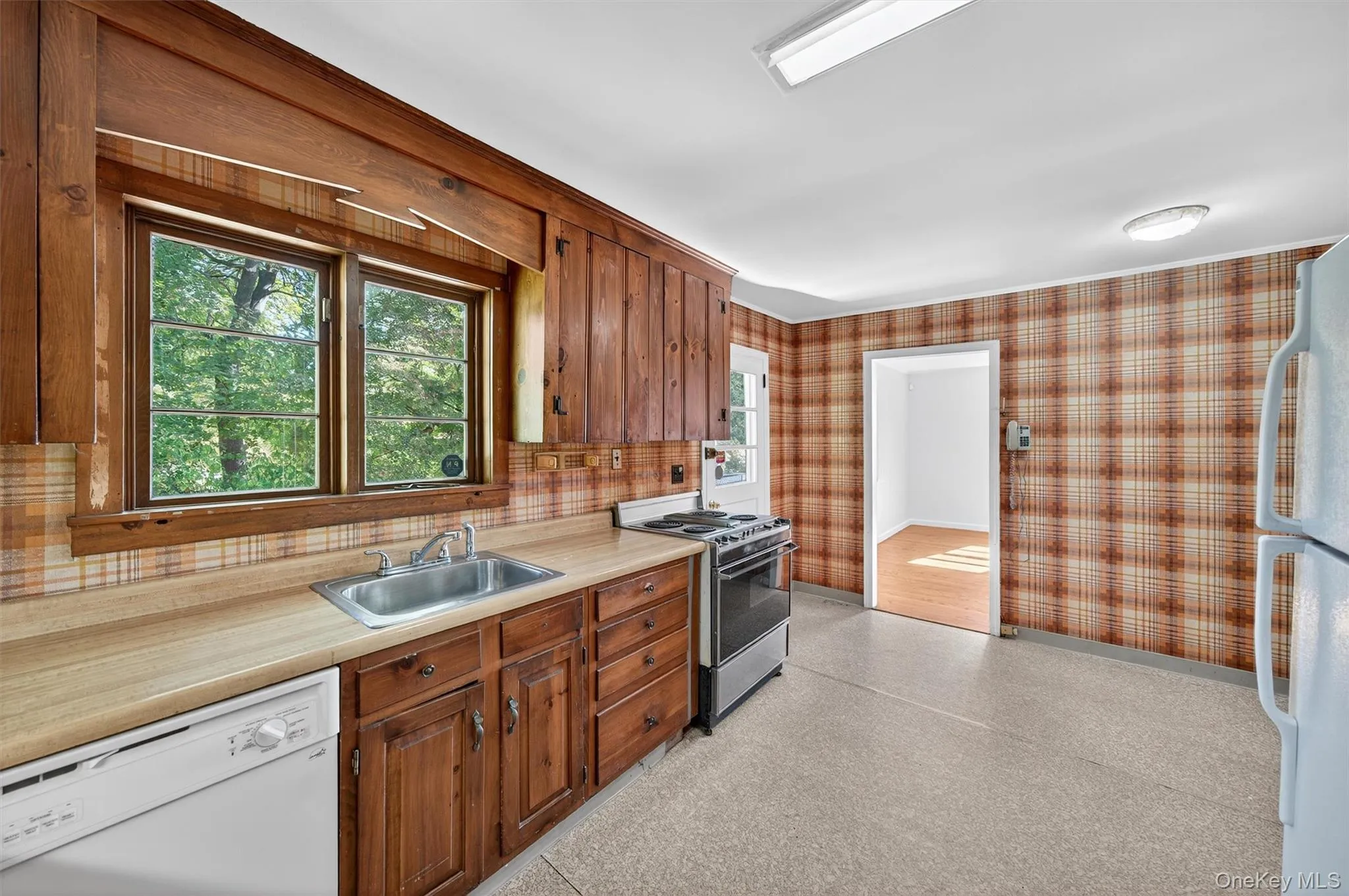 Kitchen with white appliances, light countertops, wallpapered walls, and brown cabinetry Kitchen with white appliances, light countertops, wallpapered walls, and brown cabinetry