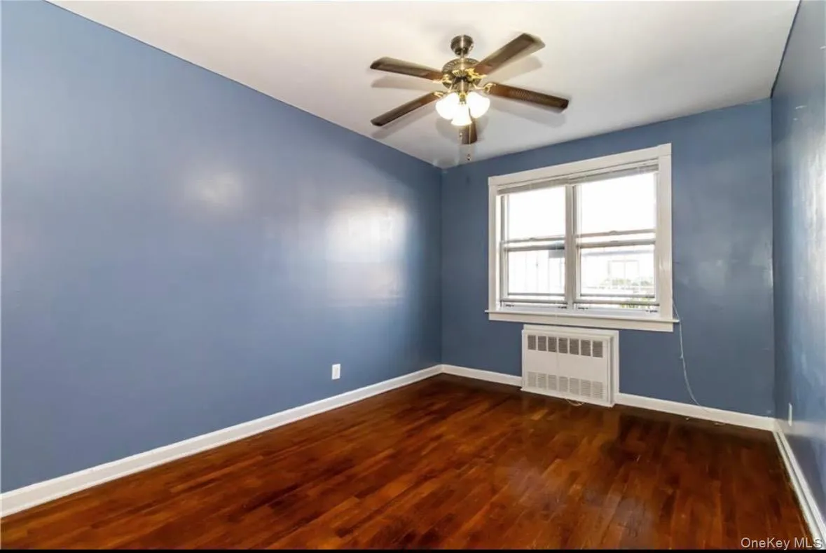 Empty room featuring dark wood-style flooring, radiator heating unit, and a ceiling fan Empty room featuring dark wood-style flooring, radiator heating unit, and a ceiling fan
