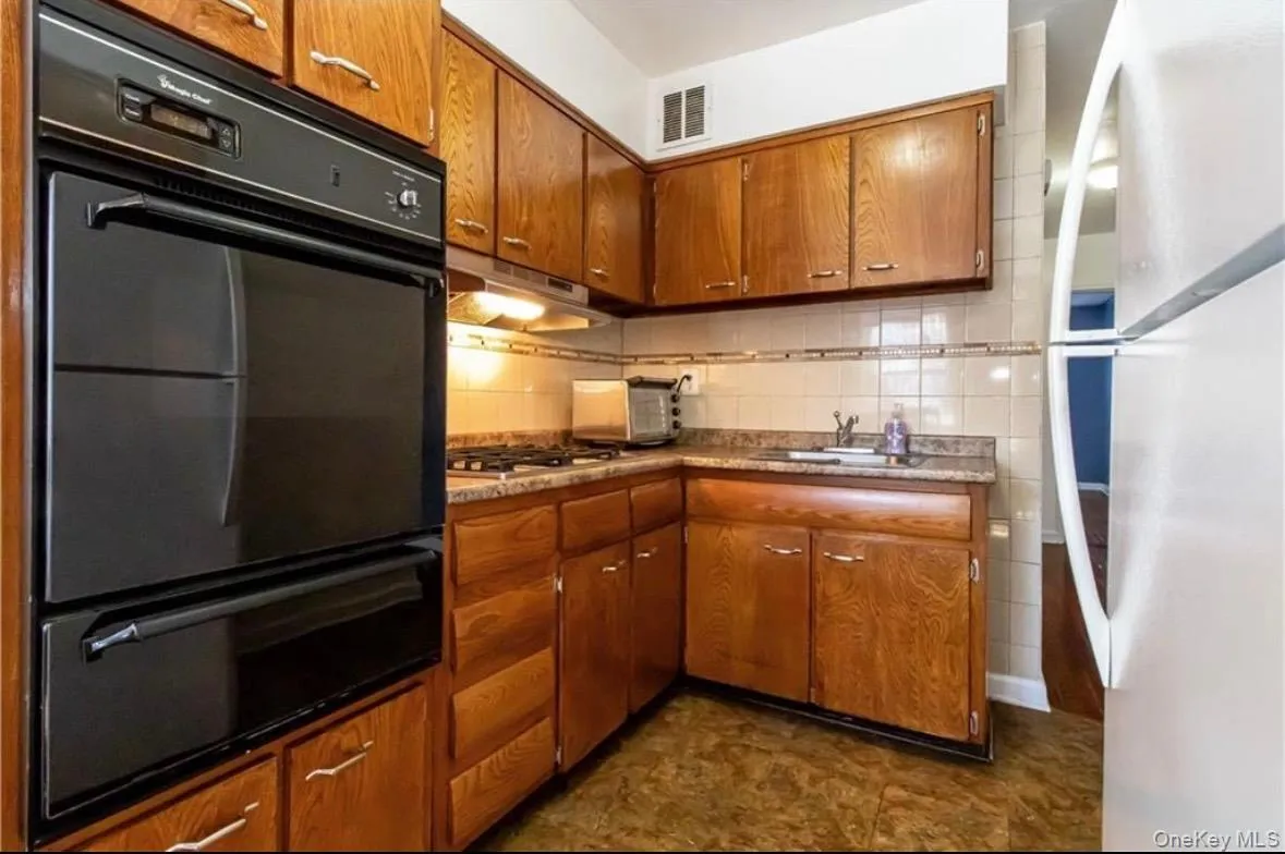 Kitchen featuring freestanding refrigerator, brown cabinetry, black oven, a warming drawer, and backsplash Kitchen featuring freestanding refrigerator, brown cabinetry, black oven, a warming drawer, and backsplash