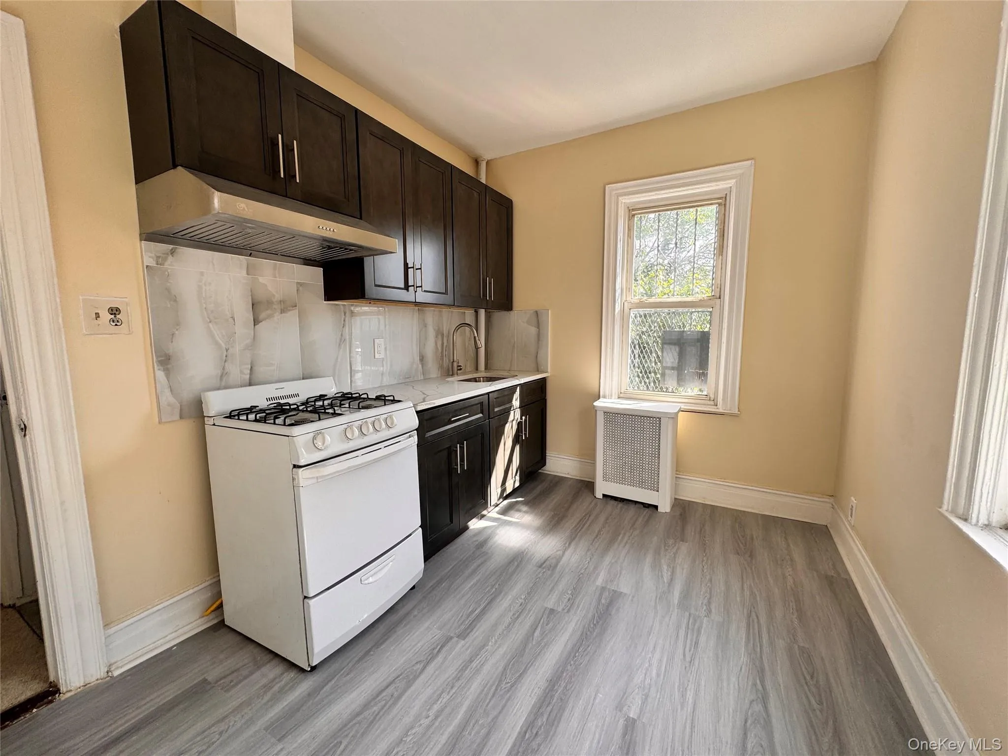 Kitchen with tasteful backsplash, white gas stove, radiator, under cabinet range hood, and light wood-style flooring Kitchen with tasteful backsplash, white gas stove, radiator, under cabinet range hood, and light wood-style flooring