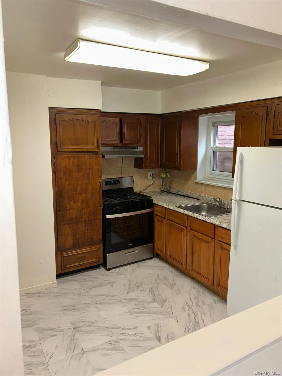 Kitchen featuring freestanding refrigerator, gas stove, backsplash, under cabinet range hood, and light marble finish flooring Kitchen featuring freestanding refrigerator, gas stove, backsplash, under cabinet range hood, and light marble finish flooring