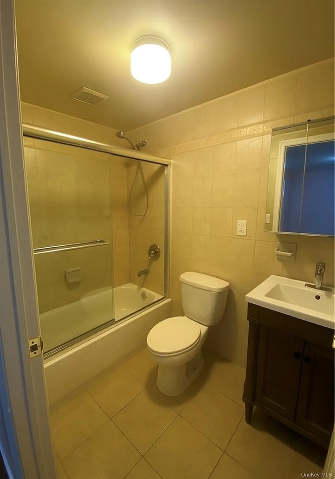 Full bathroom with tile walls, bath / shower combo with glass door, vanity, and light tile patterned floors Full bathroom with tile walls, bath / shower combo with glass door, vanity, and light tile patterned floors