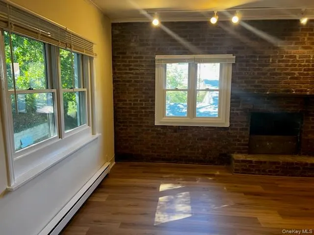 Unfurnished living room with brick wall, track lighting, a baseboard radiator, dark wood-style floors, and a brick fireplace Unfurnished living room with brick wall, track lighting, a baseboard radiator, dark wood-style floors, and a brick fireplace