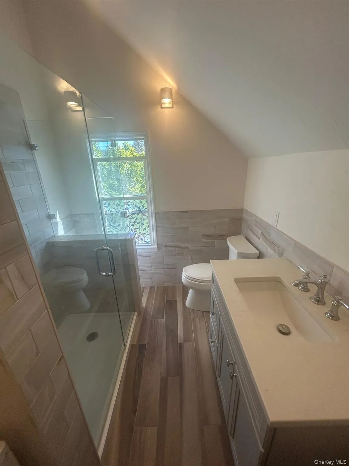 Bathroom with tile walls, a stall shower, vanity, wainscoting, and lofted ceiling Bathroom with tile walls, a stall shower, vanity, wainscoting, and lofted ceiling