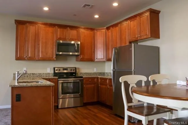 Kitchen featuring light stone countertops, brown cabinetry, stainless steel appliances, a peninsula, and dark wood finished floors Kitchen featuring light stone countertops, brown cabinetry, stainless steel appliances, a peninsula, and dark wood finished floors
