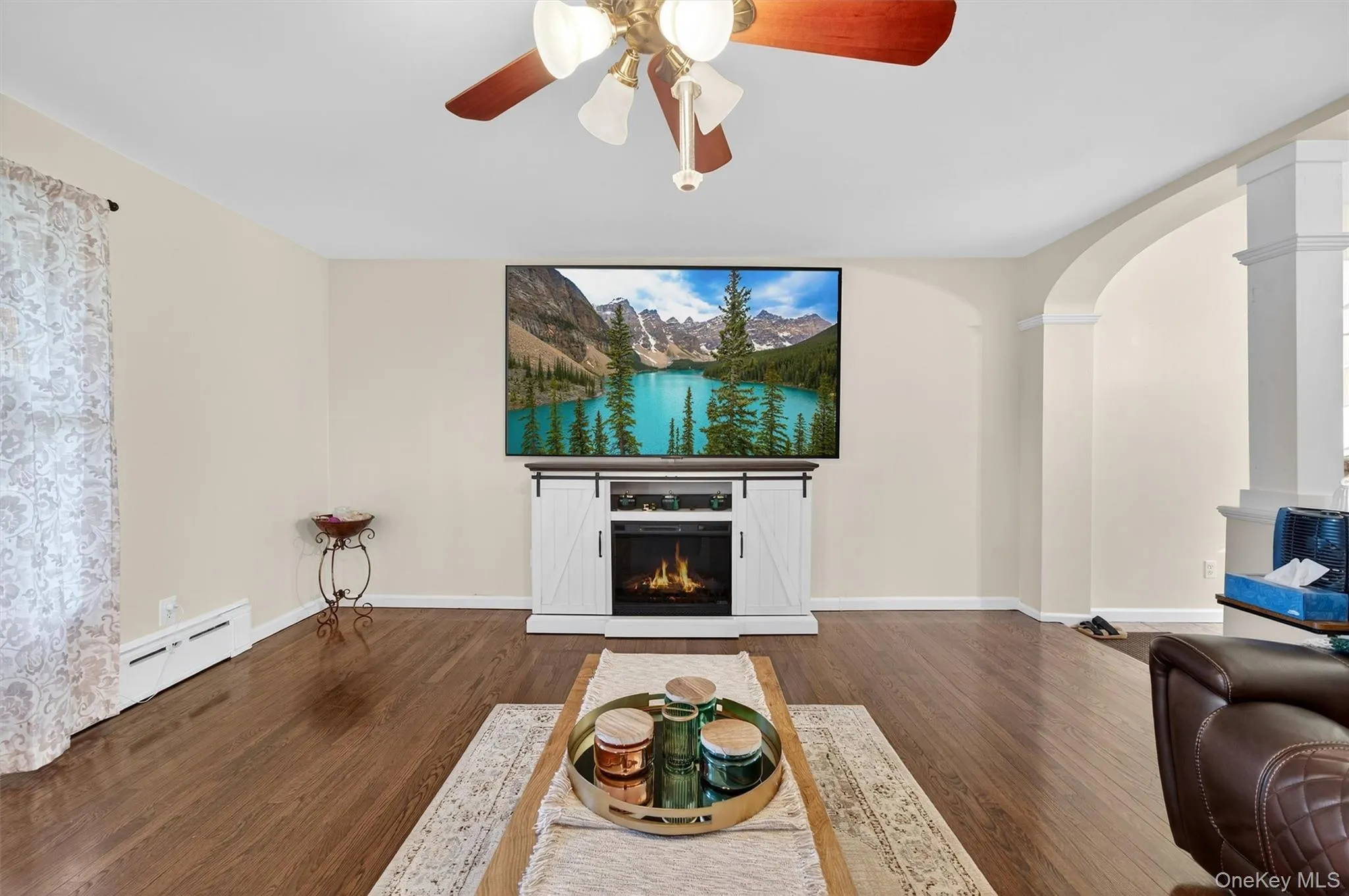 Unfurnished living room with dark wood-style flooring, a ceiling fan, a baseboard radiator, a glass covered fireplace, and arched walkways Unfurnished living room with dark wood-style flooring, a ceiling fan, a baseboard radiator, a glass covered fireplace, and arched walkways