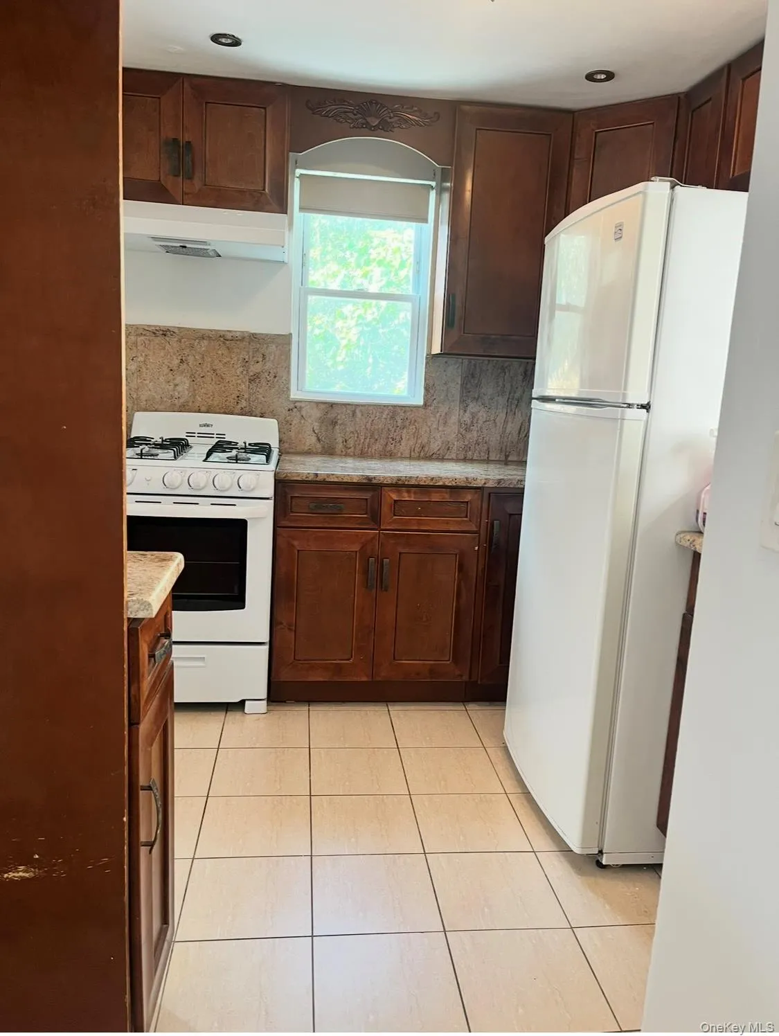 Kitchen with white appliances, light tile patterned flooring, under cabinet range hood, decorative backsplash, and light stone countertops Kitchen with white appliances, light tile patterned flooring, under cabinet range hood, decorative backsplash, and light stone countertops