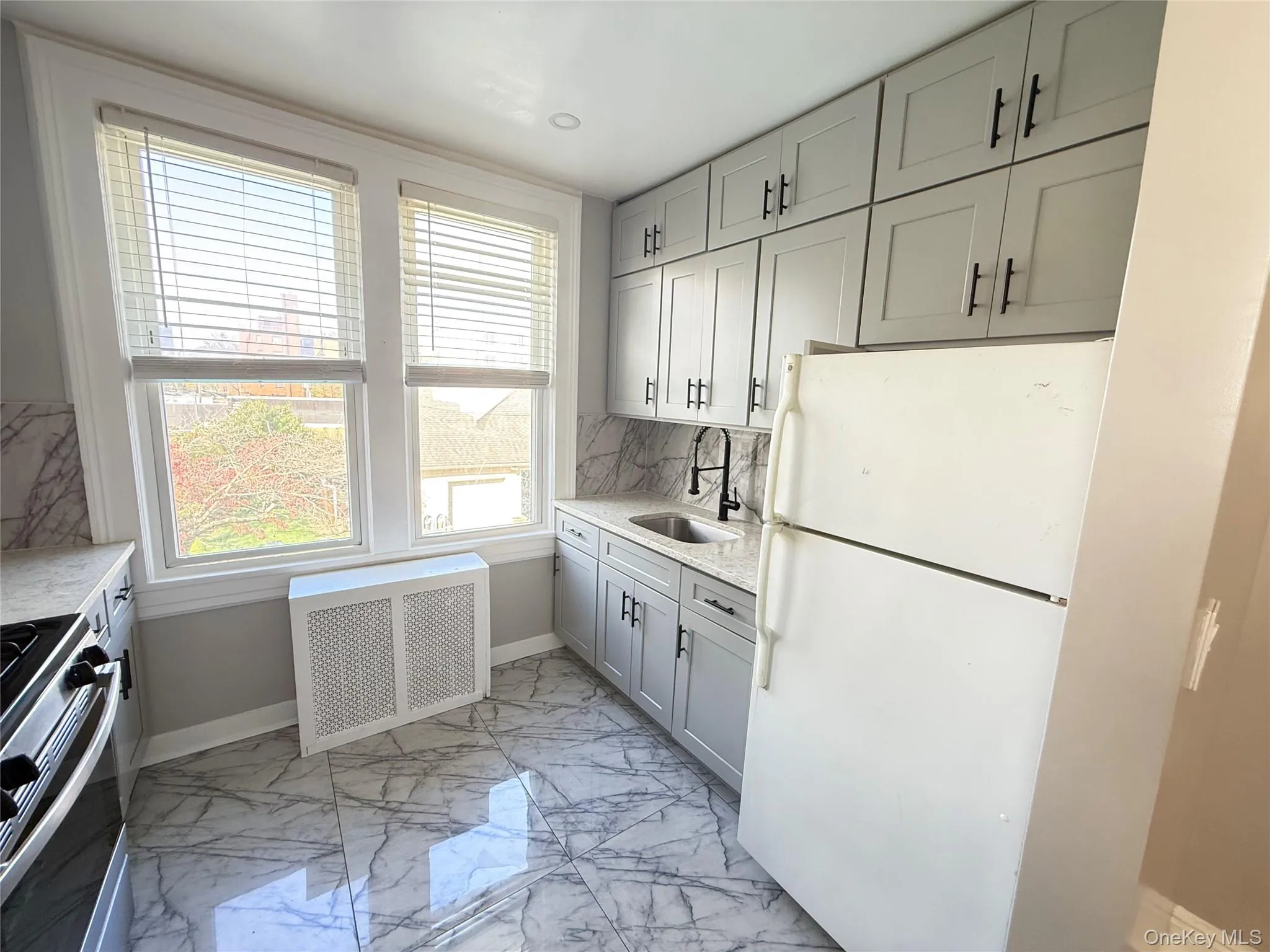 Kitchen featuring freestanding refrigerator, stainless steel gas stove, radiator, gray cabinetry, and light marble finish flooring Kitchen featuring freestanding refrigerator, stainless steel gas stove, radiator, gray cabinetry, and light marble finish flooring