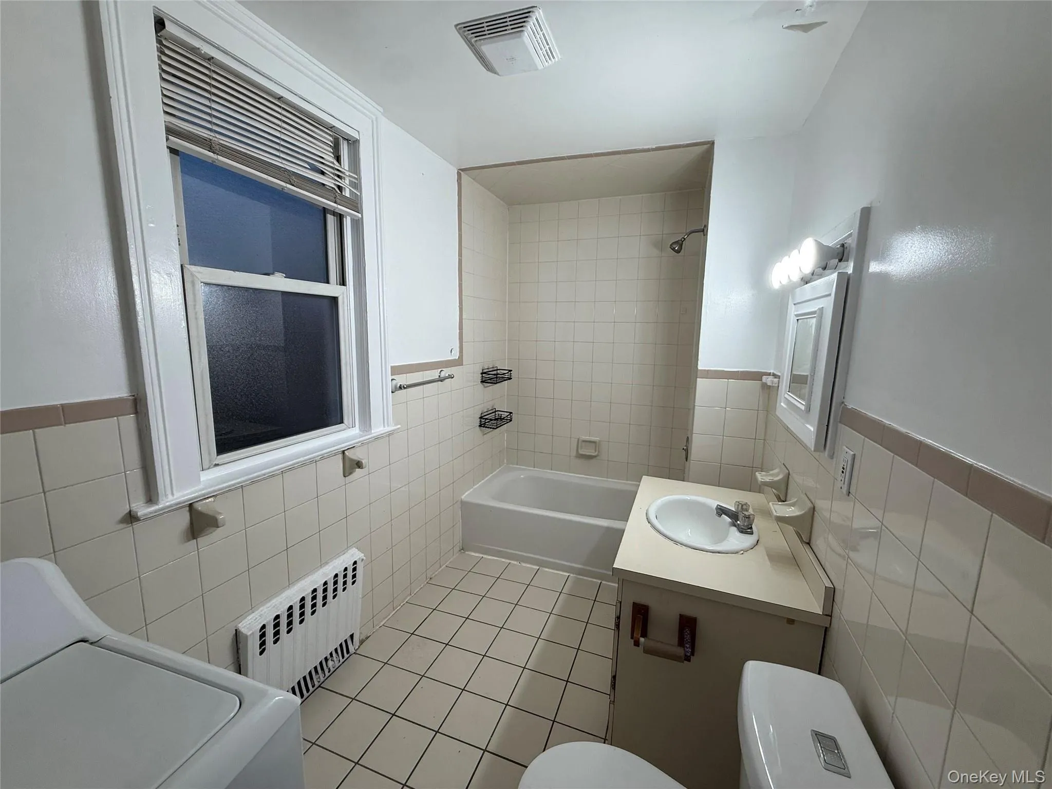 Bathroom featuring tile walls, light tile patterned floors, washer / clothes dryer, radiator heating unit, and a wainscoted wall Bathroom featuring tile walls, light tile patterned floors, washer / clothes dryer, radiator heating unit, and a wainscoted wall