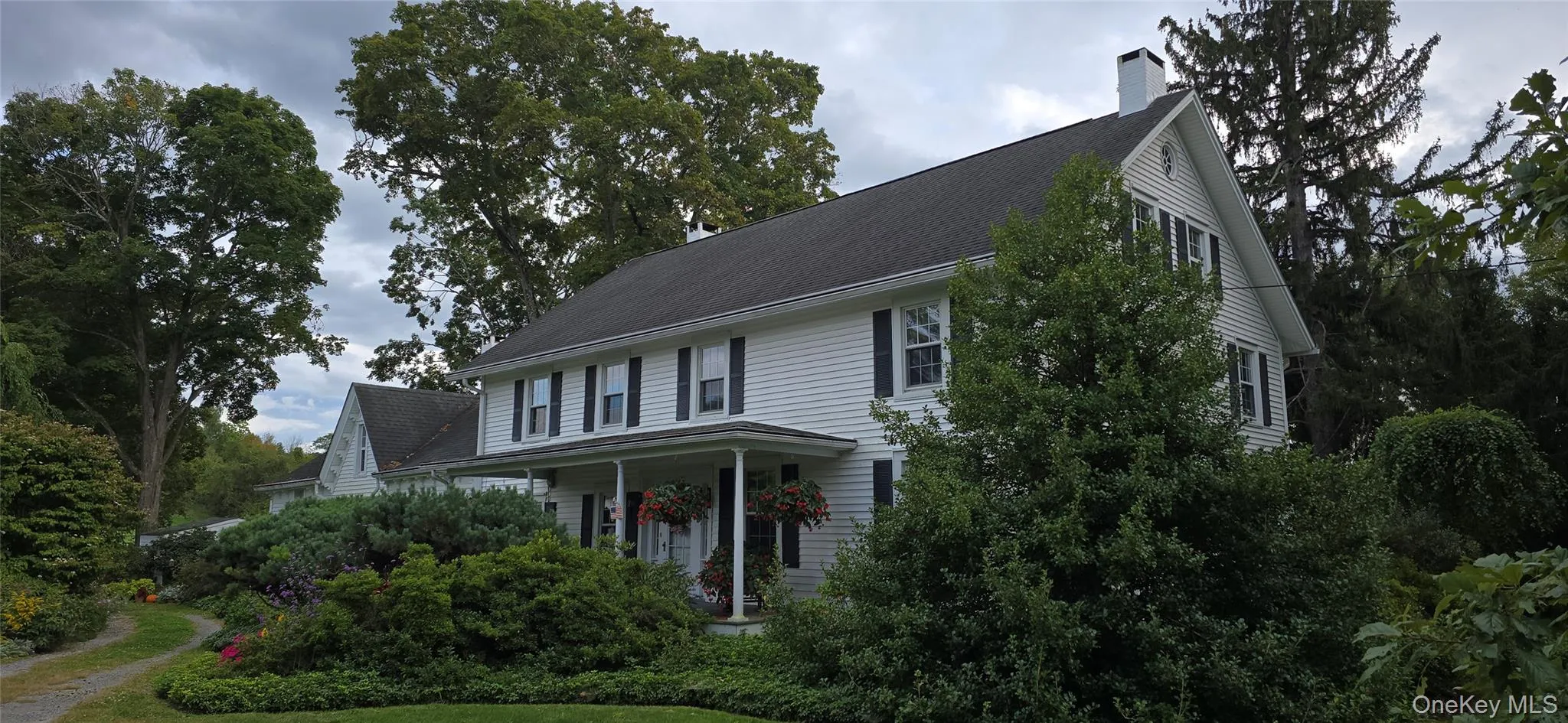 View of front of property featuring a chimney, a porch, view of scattered trees, and roof with shingles View of front of property featuring a chimney, a porch, view of scattered trees, and roof with shingles