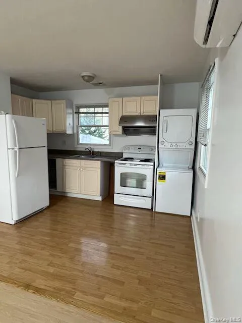 Kitchen with white appliances, dark countertops, light wood-style flooring, stacked washing machine and dryer, and a wall unit AC Kitchen with white appliances, dark countertops, light wood-style flooring, stacked washing machine and dryer, and a wall unit AC