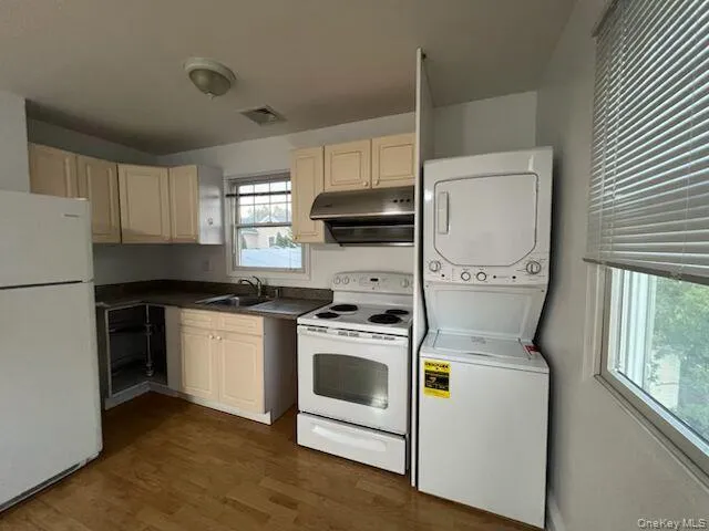 Kitchen featuring white appliances, dark wood-style floors, stacked washer / dryer, dark countertops, and under cabinet range hood Kitchen featuring white appliances, dark wood-style floors, stacked washer / dryer, dark countertops, and under cabinet range hood