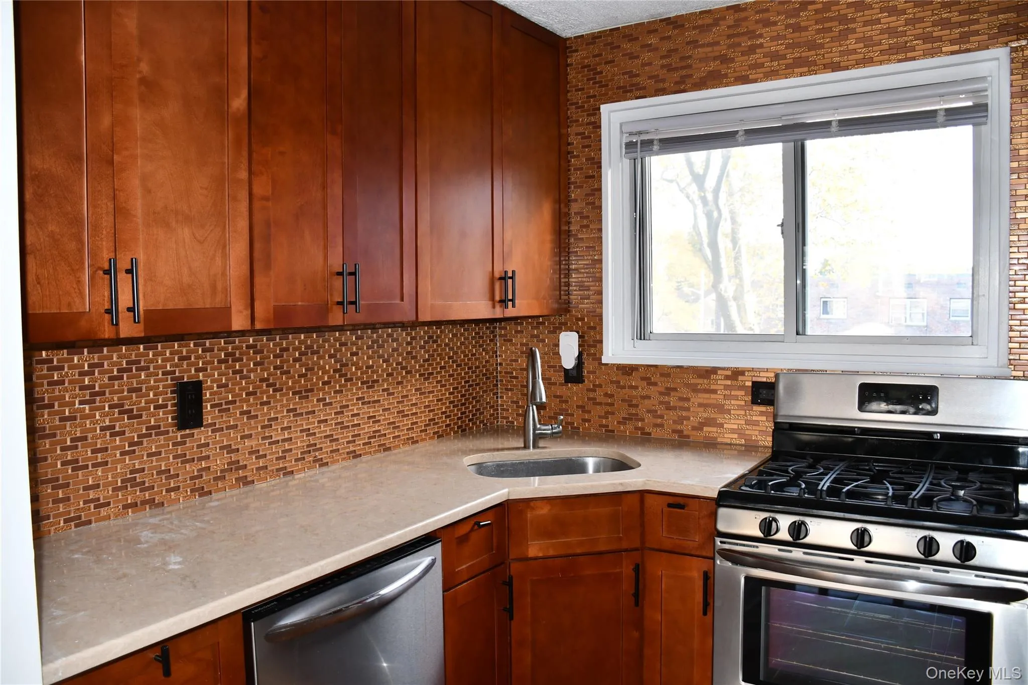 Kitchen featuring stainless steel appliances, decorative backsplash, light stone countertops, and brown cabinetry Kitchen featuring stainless steel appliances, decorative backsplash, light stone countertops, and brown cabinetry