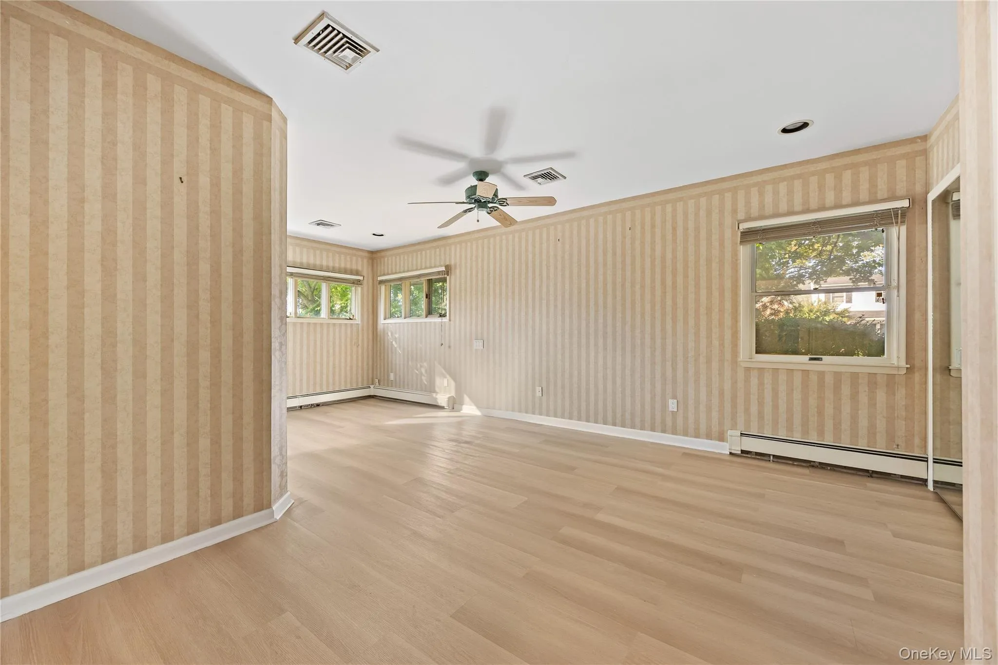 Empty room featuring light wood-type flooring, wallpapered walls, a baseboard radiator, plenty of natural light, and a ceiling fan Empty room featuring light wood-type flooring, wallpapered walls, a baseboard radiator, plenty of natural light, and a ceiling fan