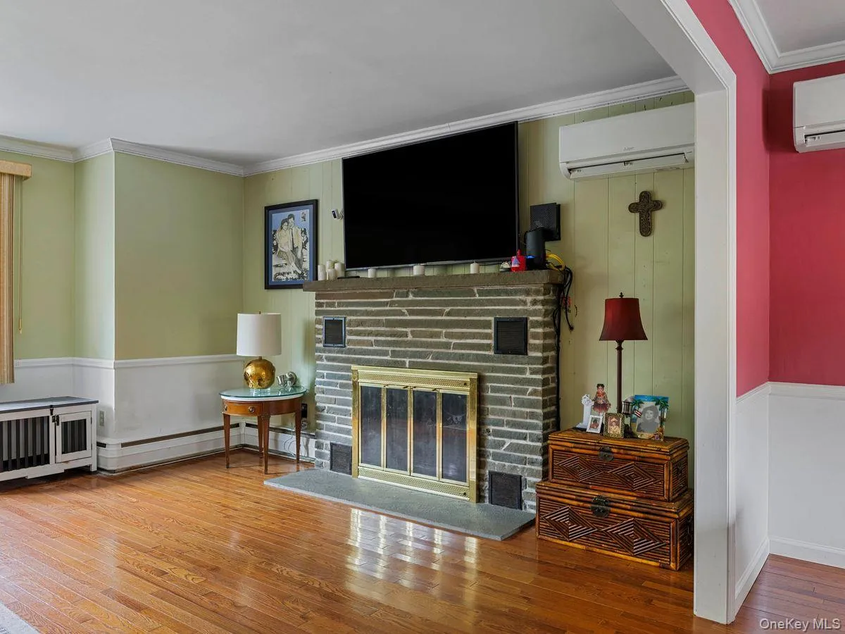 Living room with ornamental molding, hardwood / wood-style flooring, a wall mounted air conditioner, a fireplace, and baseboard heating Living room with ornamental molding, hardwood / wood-style flooring, a wall mounted air conditioner, a fireplace, and baseboard heating