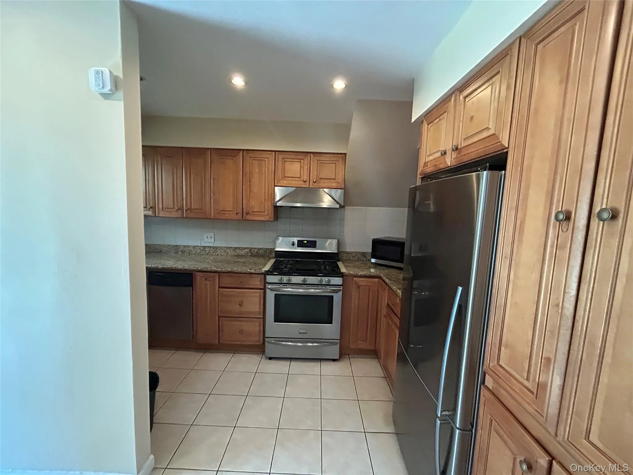Kitchen featuring appliances with stainless steel finishes, backsplash, brown cabinetry, light tile patterned floors, and dark stone counters Kitchen featuring appliances with stainless steel finishes, backsplash, brown cabinetry, light tile patterned floors, and dark stone counters