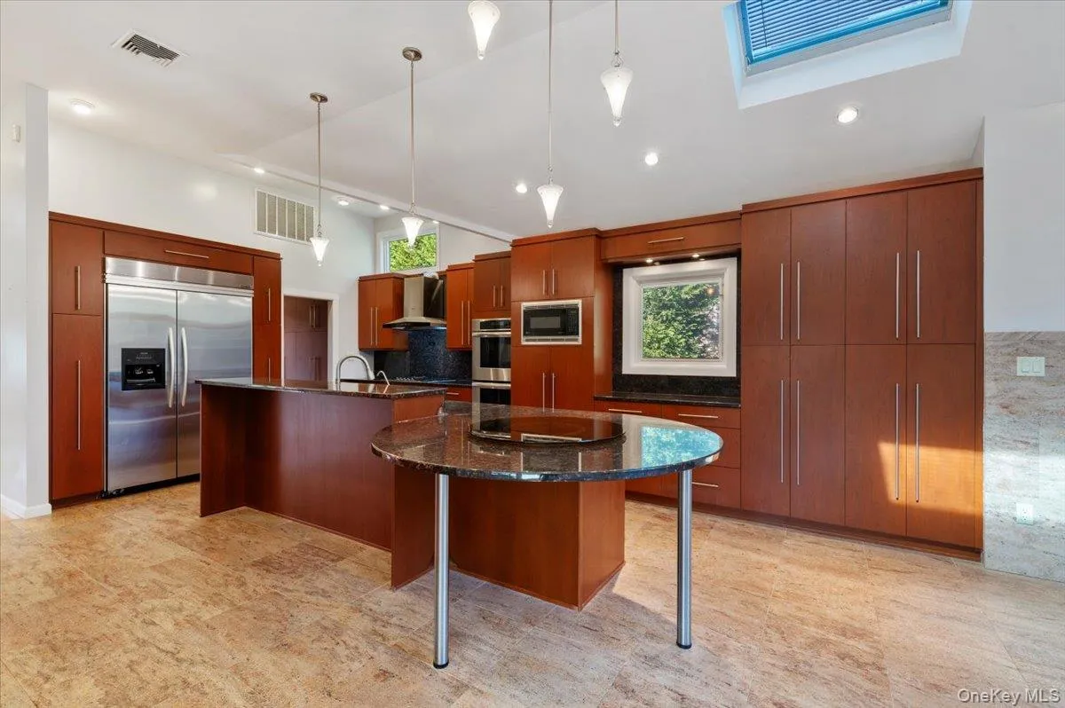 Kitchen with dark stone countertops, pendant lighting, built in appliances, decorative backsplash, and wall chimney range hood Kitchen with dark stone countertops, pendant lighting, built in appliances, decorative backsplash, and wall chimney range hood