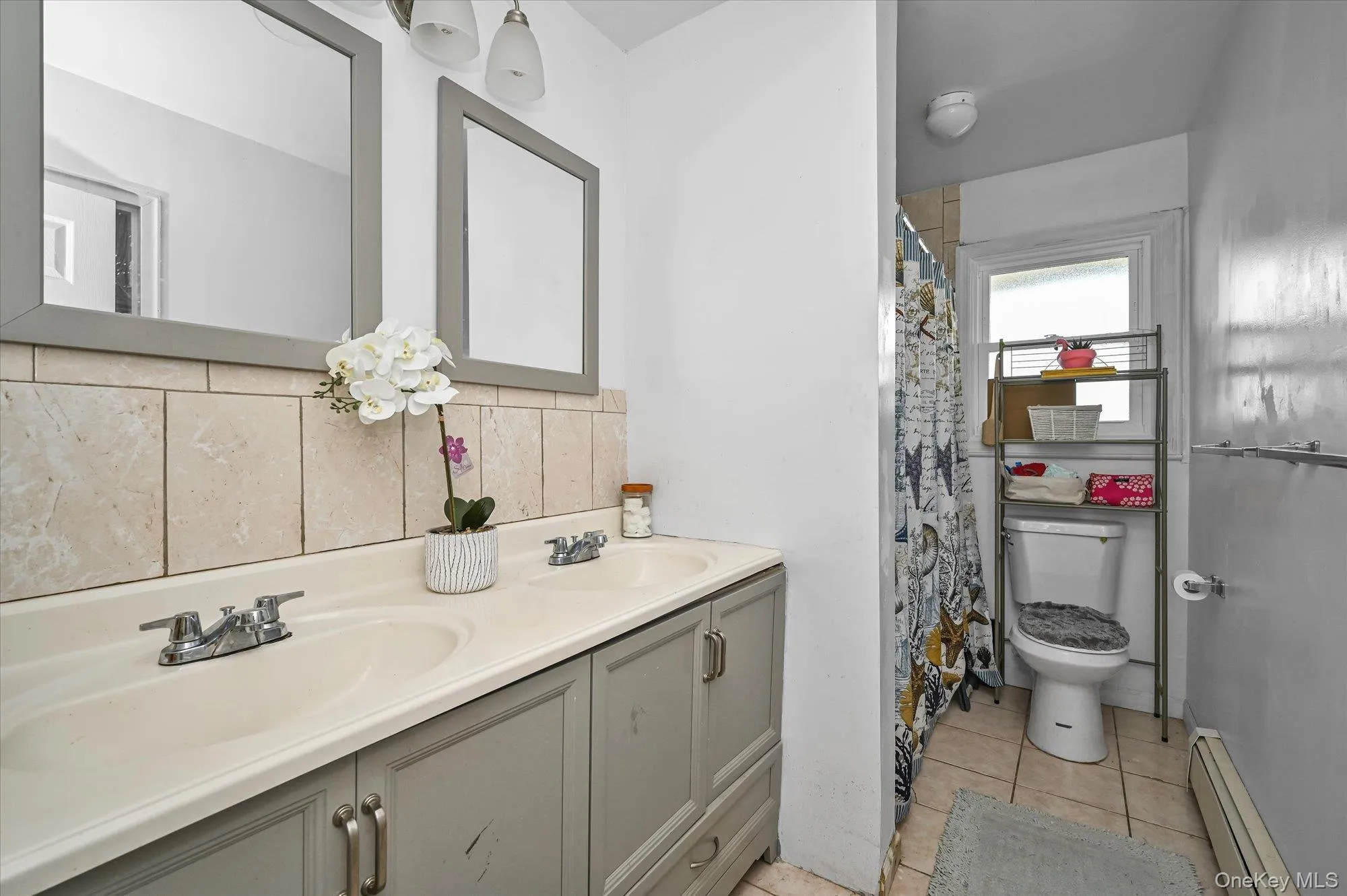 Full bathroom featuring baseboard heating, light tile patterned flooring, a shower with curtain, double vanity, and backsplash Full bathroom featuring baseboard heating, light tile patterned flooring, a shower with curtain, double vanity, and backsplash