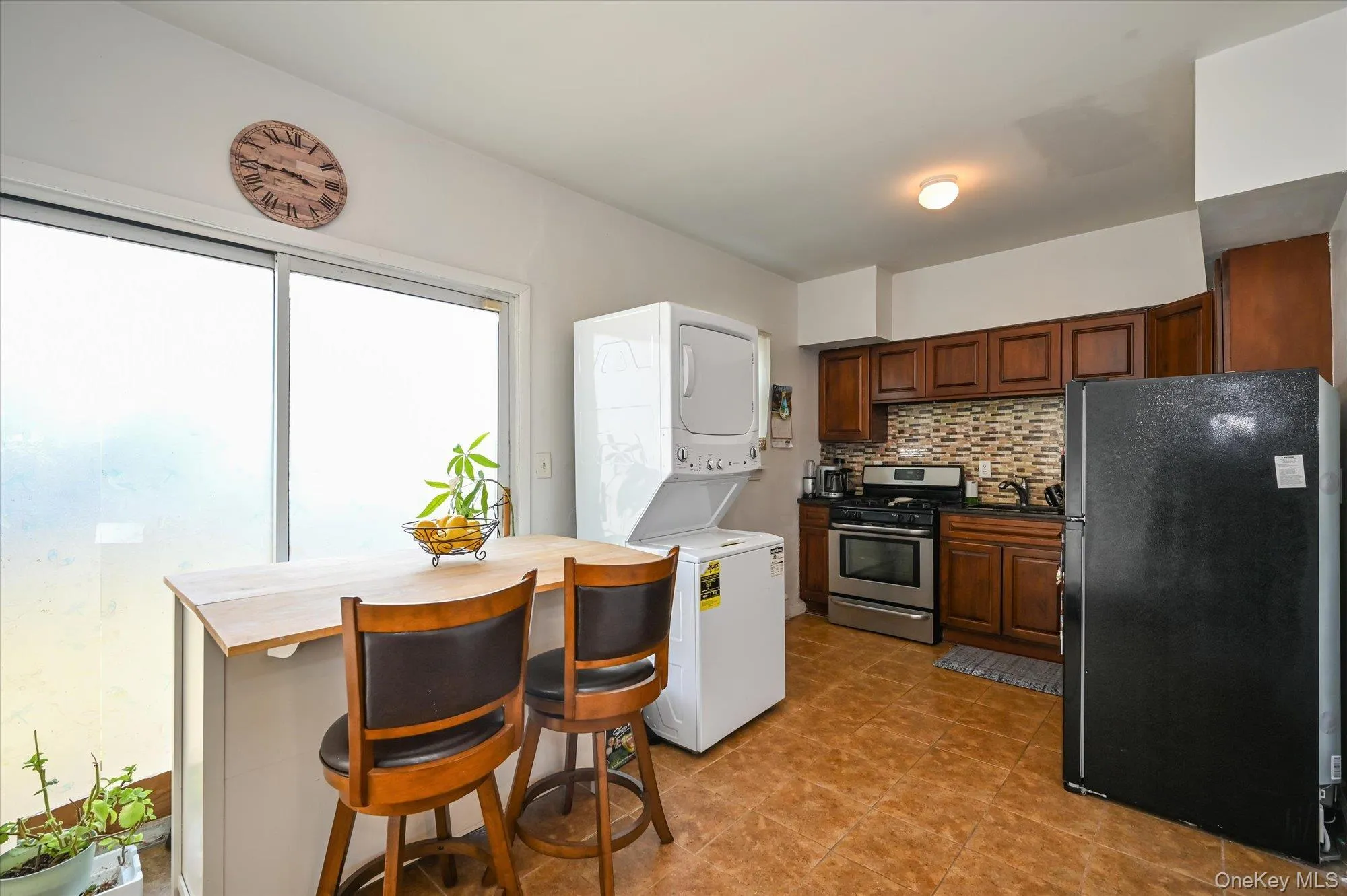 Kitchen with freestanding refrigerator, decorative backsplash, stainless steel gas stove, stacked washer / dryer, and dark countertops Kitchen with freestanding refrigerator, decorative backsplash, stainless steel gas stove, stacked washer / dryer, and dark countertops
