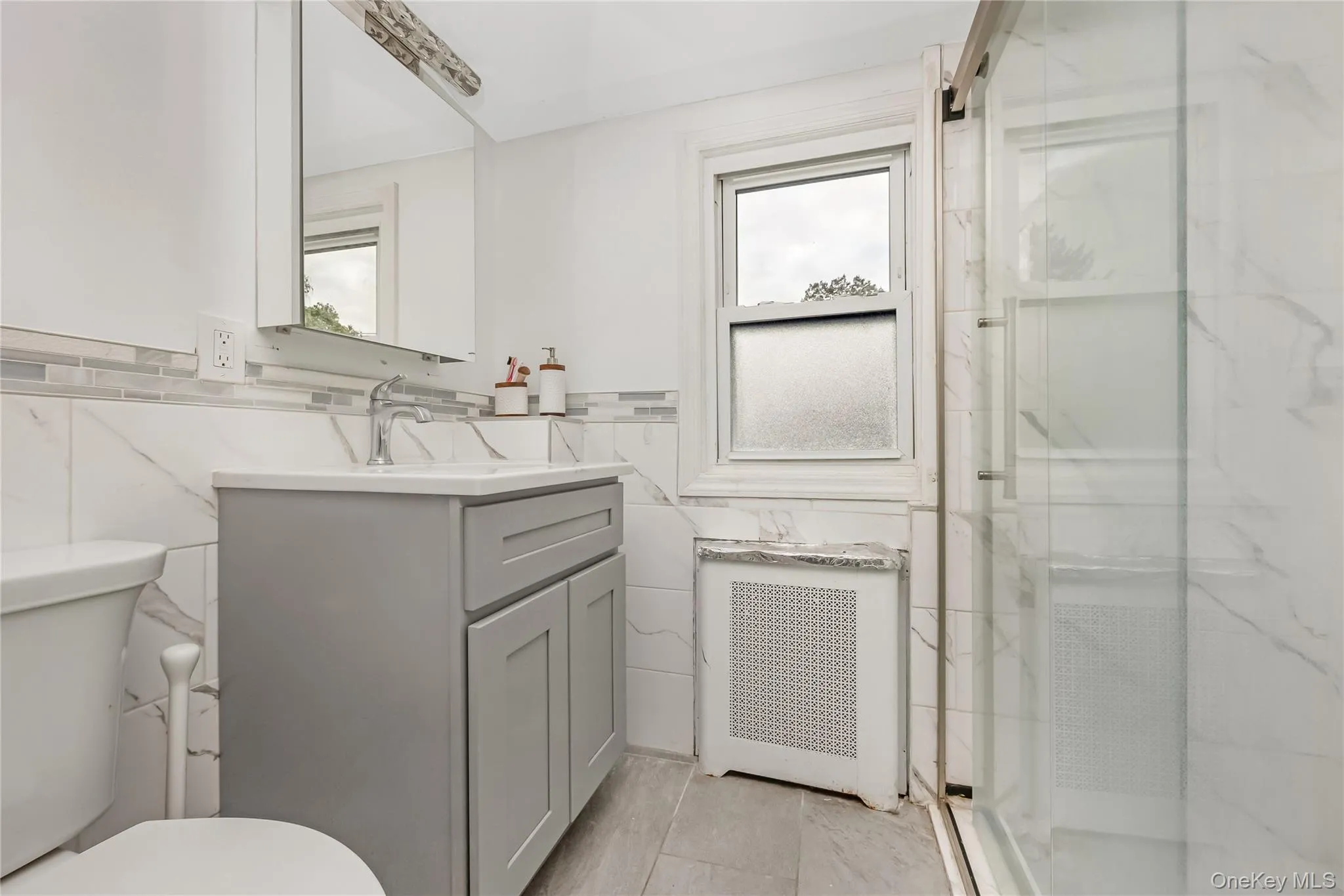 Full bathroom featuring tile walls, radiator, vanity, a marble finish shower, and wainscoting Full bathroom featuring tile walls, radiator, vanity, a marble finish shower, and wainscoting