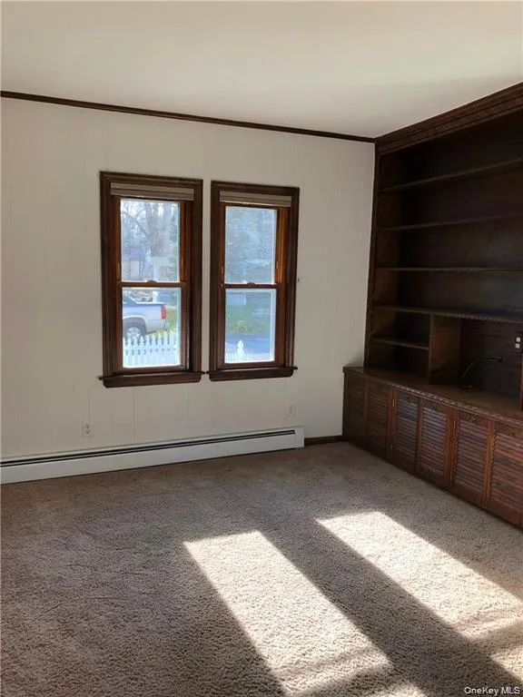 Unfurnished living room featuring a baseboard heating unit, carpet flooring, wood walls, crown molding, and built in shelves Unfurnished living room featuring a baseboard heating unit, carpet flooring, wood walls, crown molding, and built in shelves