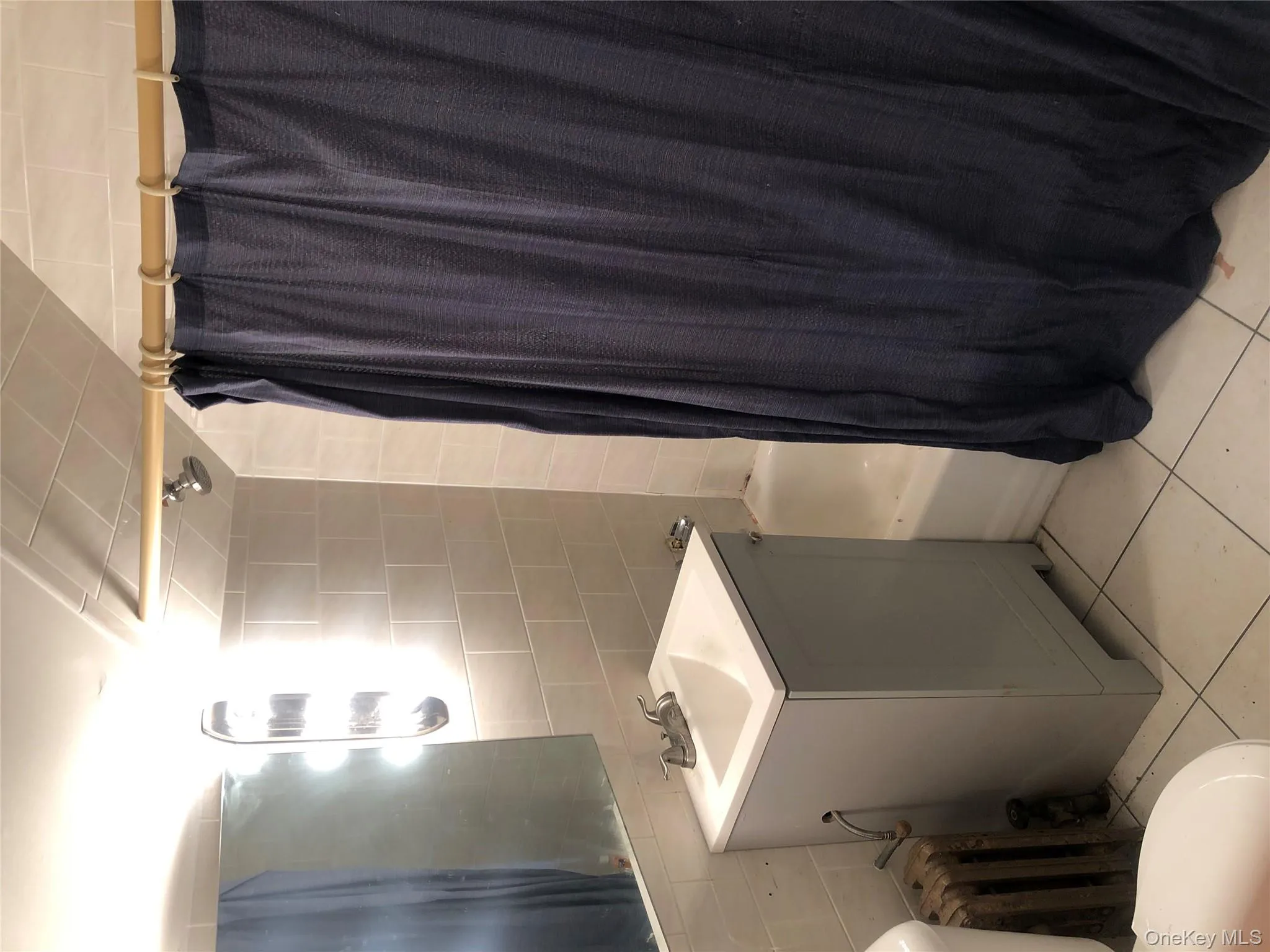 Full bath featuring tile walls, shower / tub combo, light tile patterned floors, vanity, and radiator heating unit Full bath featuring tile walls, shower / tub combo, light tile patterned floors, vanity, and radiator heating unit