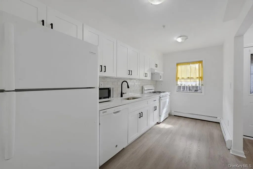 Kitchen featuring white appliances, white cabinets, backsplash, light wood-style flooring, and a baseboard radiator Kitchen featuring white appliances, white cabinets, backsplash, light wood-style flooring, and a baseboard radiator