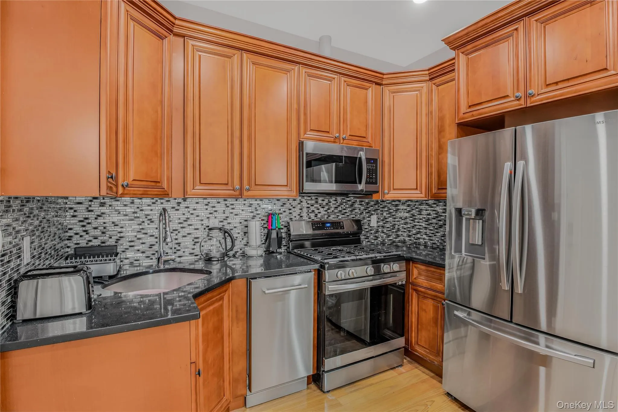Kitchen with stainless steel appliances, dark stone countertops, light wood-type flooring, brown cabinetry, and decorative backsplash Kitchen with stainless steel appliances, dark stone countertops, light wood-type flooring, brown cabinetry, and decorative backsplash