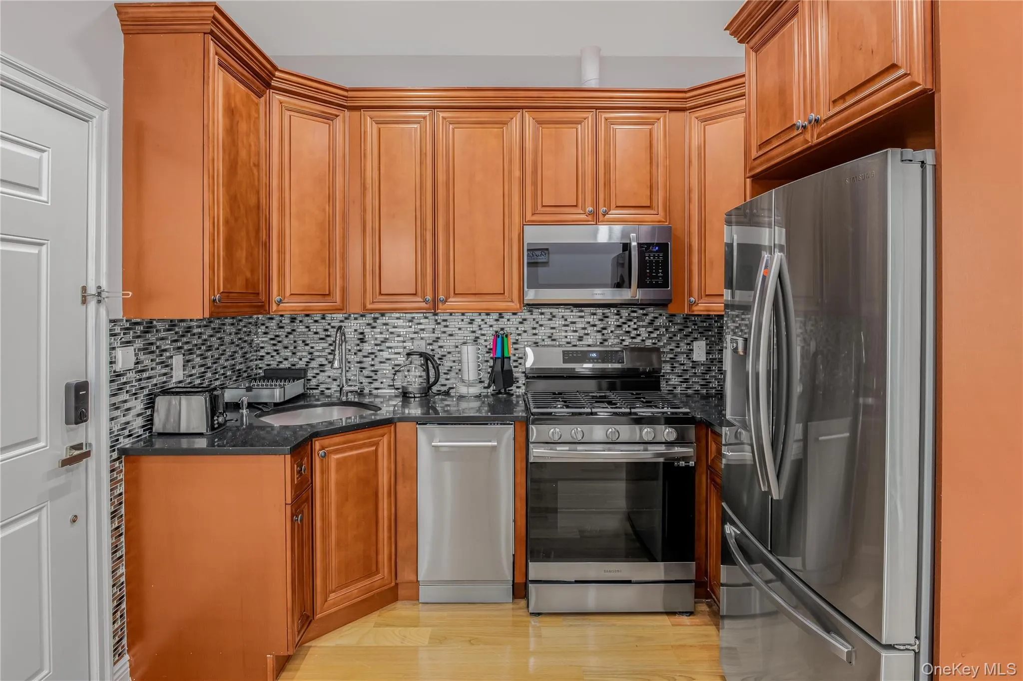 Kitchen with appliances with stainless steel finishes, dark stone counters, tasteful backsplash, brown cabinets, and light wood-style floors Kitchen with appliances with stainless steel finishes, dark stone counters, tasteful backsplash, brown cabinets, and light wood-style floors