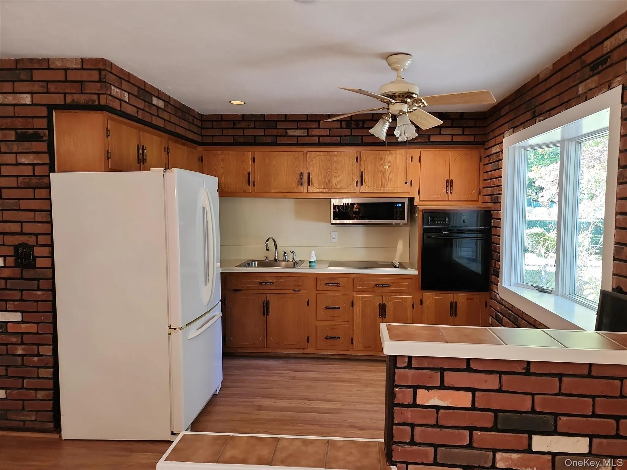 Kitchen featuring brick wall, black appliances, brown cabinetry, tile countertops, and light wood-type flooring Kitchen featuring brick wall, black appliances, brown cabinetry, tile countertops, and light wood-type flooring