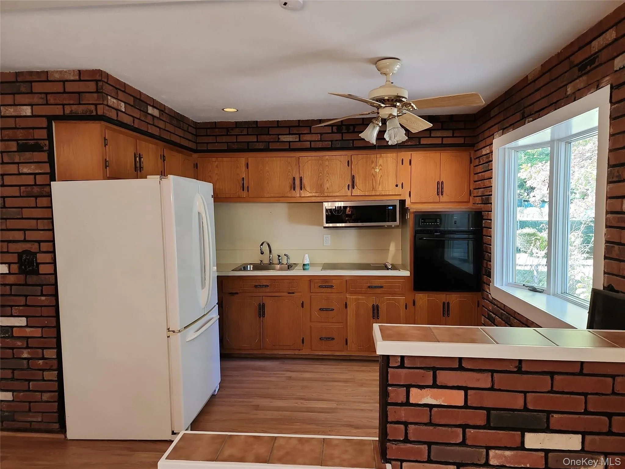 Kitchen with brick wall, black appliances, tile countertops, brown cabinetry, and light wood-type flooring Kitchen with brick wall, black appliances, tile countertops, brown cabinetry, and light wood-type flooring