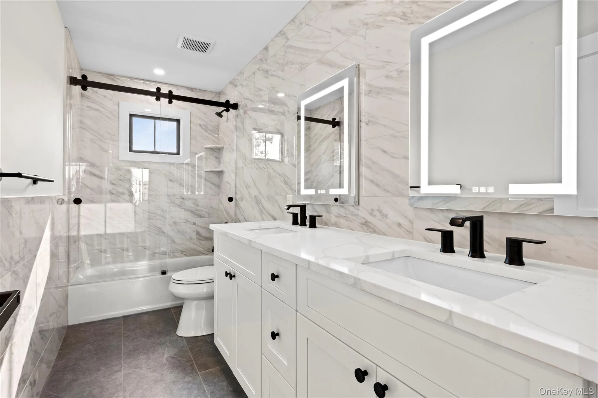 Full bath with tile walls, shower / bath combination with glass door, double vanity, and dark tile patterned flooring Full bath with tile walls, shower / bath combination with glass door, double vanity, and dark tile patterned flooring