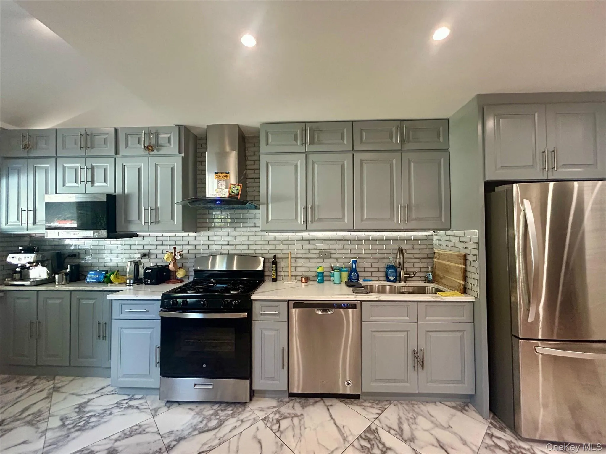 Kitchen with appliances with stainless steel finishes, light marble finish floors, wall chimney exhaust hood, gray cabinetry, and recessed lighting Kitchen with appliances with stainless steel finishes, light marble finish floors, wall chimney exhaust hood, gray cabinetry, and recessed lighting