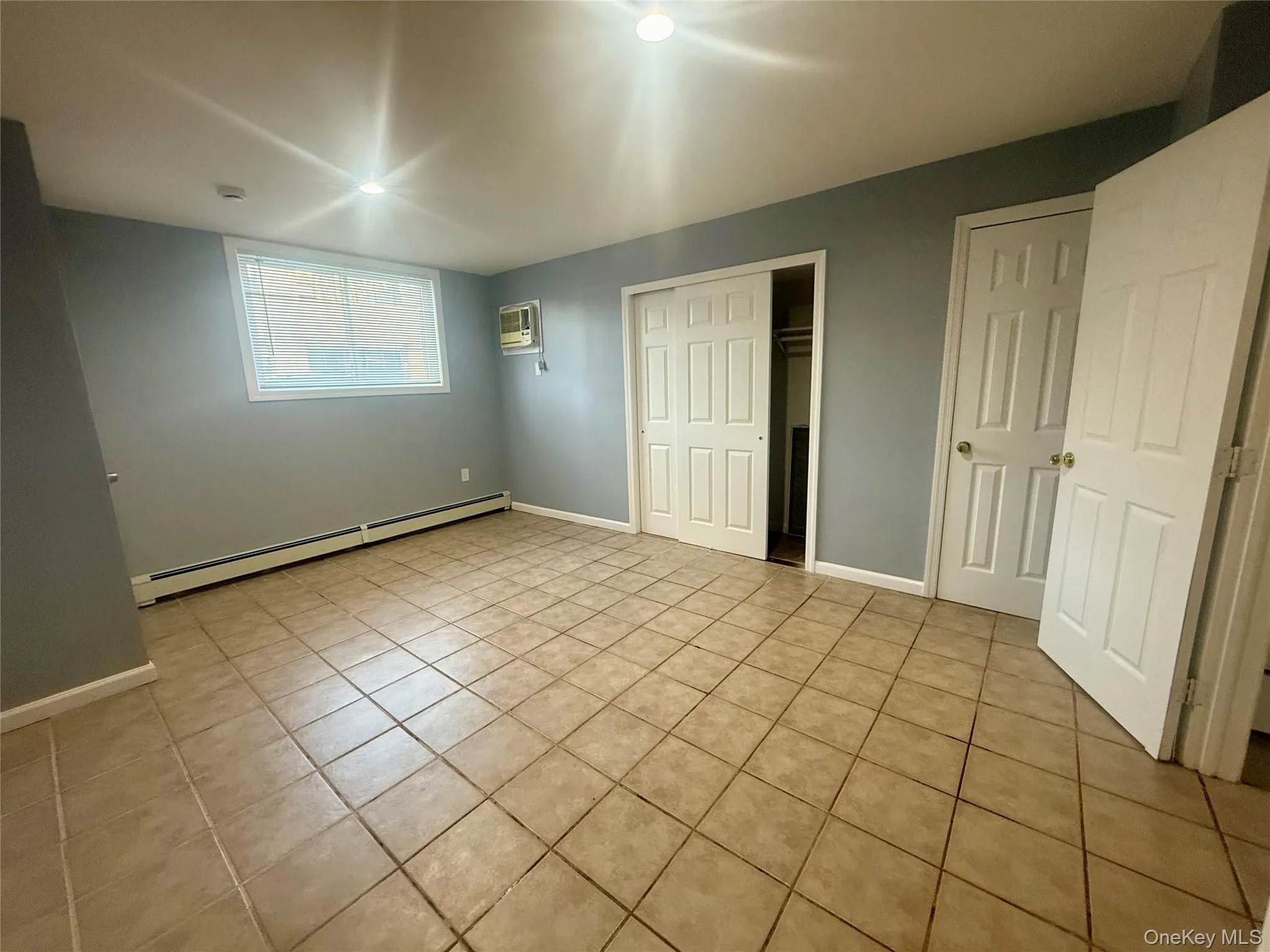 Unfurnished bedroom featuring light tile patterned floors, a baseboard radiator, a closet, and a wall mounted AC Unfurnished bedroom featuring light tile patterned floors, a baseboard radiator, a closet, and a wall mounted AC