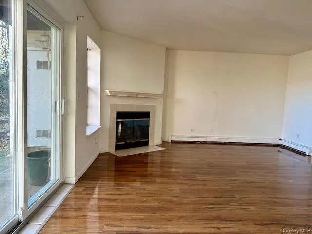 Unfurnished living room with wood finished floors, healthy amount of natural light, a fireplace, and baseboard heating Unfurnished living room with wood finished floors, healthy amount of natural light, a fireplace, and baseboard heating