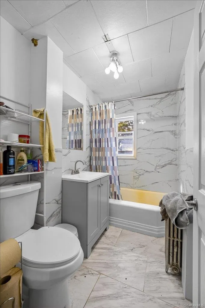 Bathroom featuring light marble finish flooring, vanity, shower / bathtub combination with curtain, and tile walls Bathroom featuring light marble finish flooring, vanity, shower / bathtub combination with curtain, and tile walls