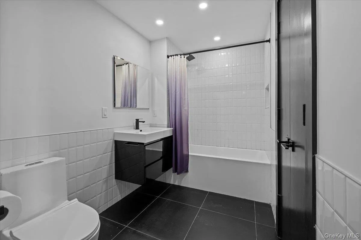 Bathroom featuring a wainscoted wall, tile walls, dark tile patterned floors, vanity, and shower / bathtub combination with curtain Bathroom featuring a wainscoted wall, tile walls, dark tile patterned floors, vanity, and shower / bathtub combination with curtain