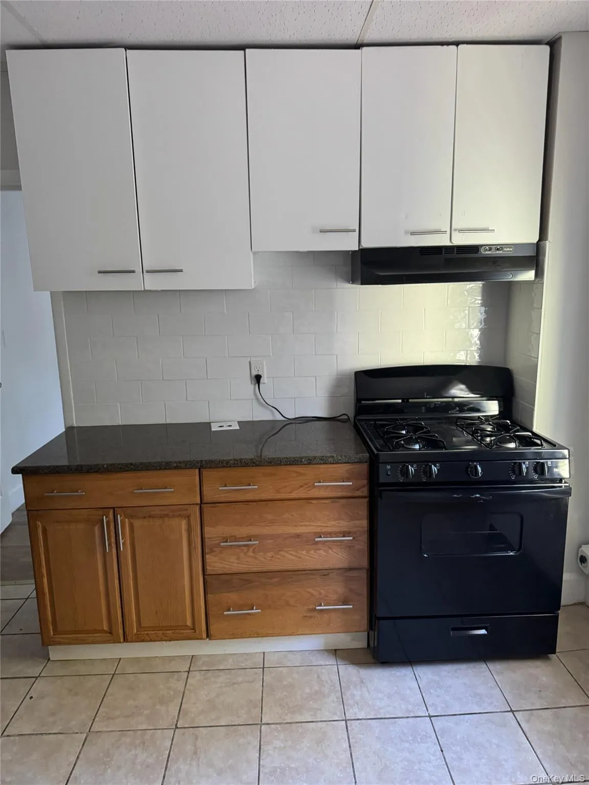 Kitchen featuring black range with gas cooktop, light tile patterned flooring, under cabinet range hood, decorative backsplash, and dark stone countertops Kitchen featuring black range with gas cooktop, light tile patterned flooring, under cabinet range hood, decorative backsplash, and dark stone countertops