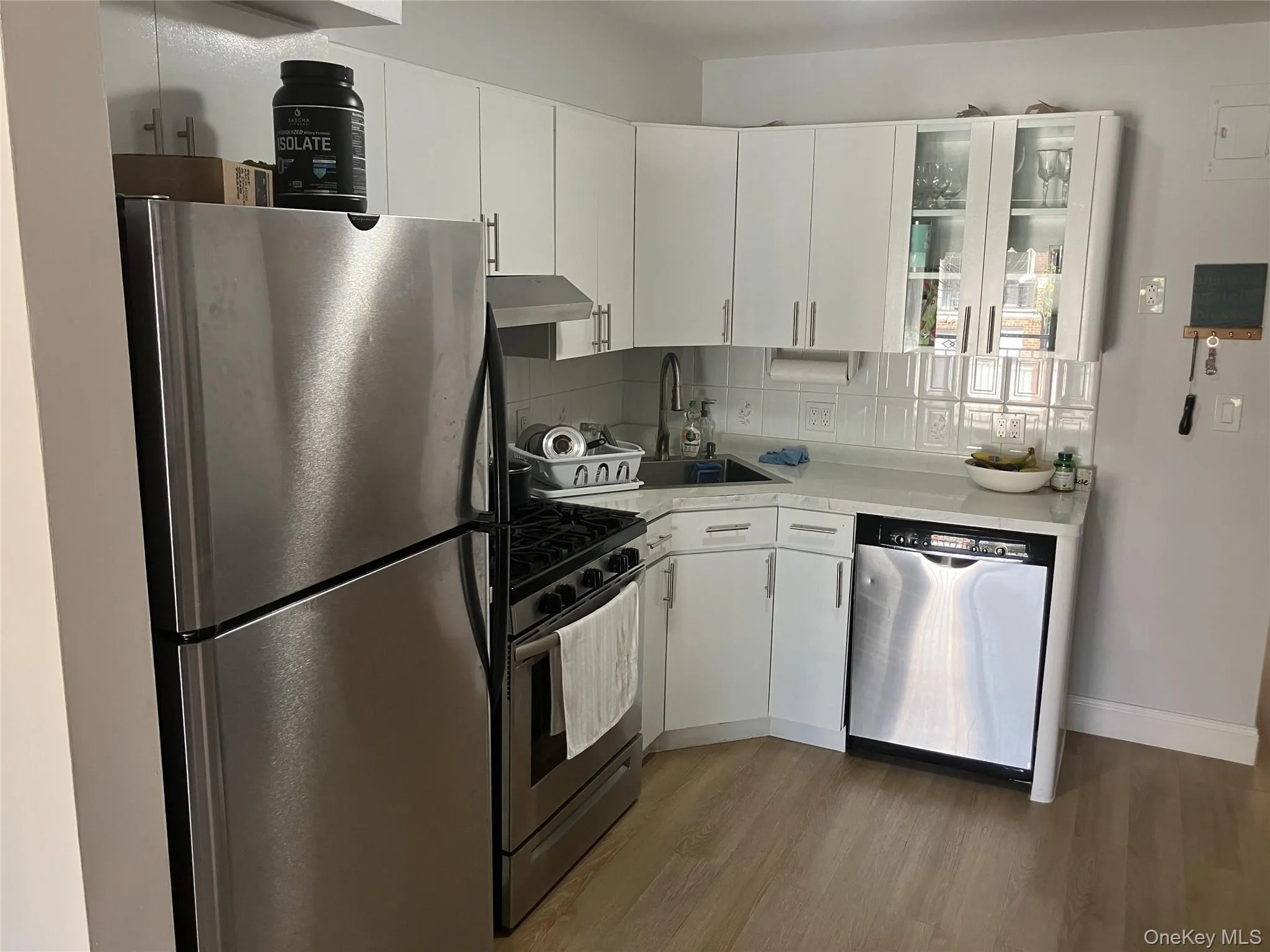 Kitchen featuring appliances with stainless steel finishes, glass insert cabinets, white cabinetry, dark wood-type flooring, and under cabinet range hood Kitchen featuring appliances with stainless steel finishes, glass insert cabinets, white cabinetry, dark wood-type flooring, and under cabinet range hood