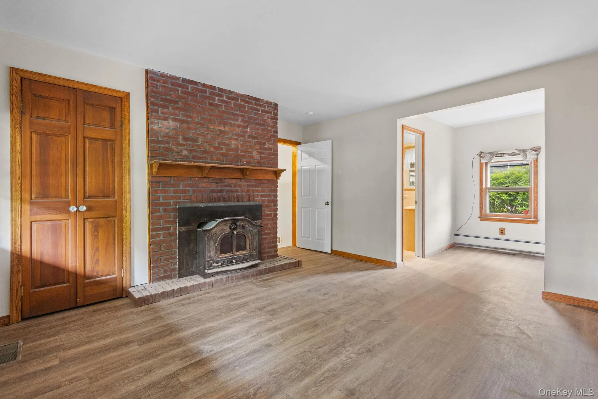 Unfurnished living room with baseboard heating, wood finished floors, baseboards, and a brick fireplace Unfurnished living room with baseboard heating, wood finished floors, baseboards, and a brick fireplace