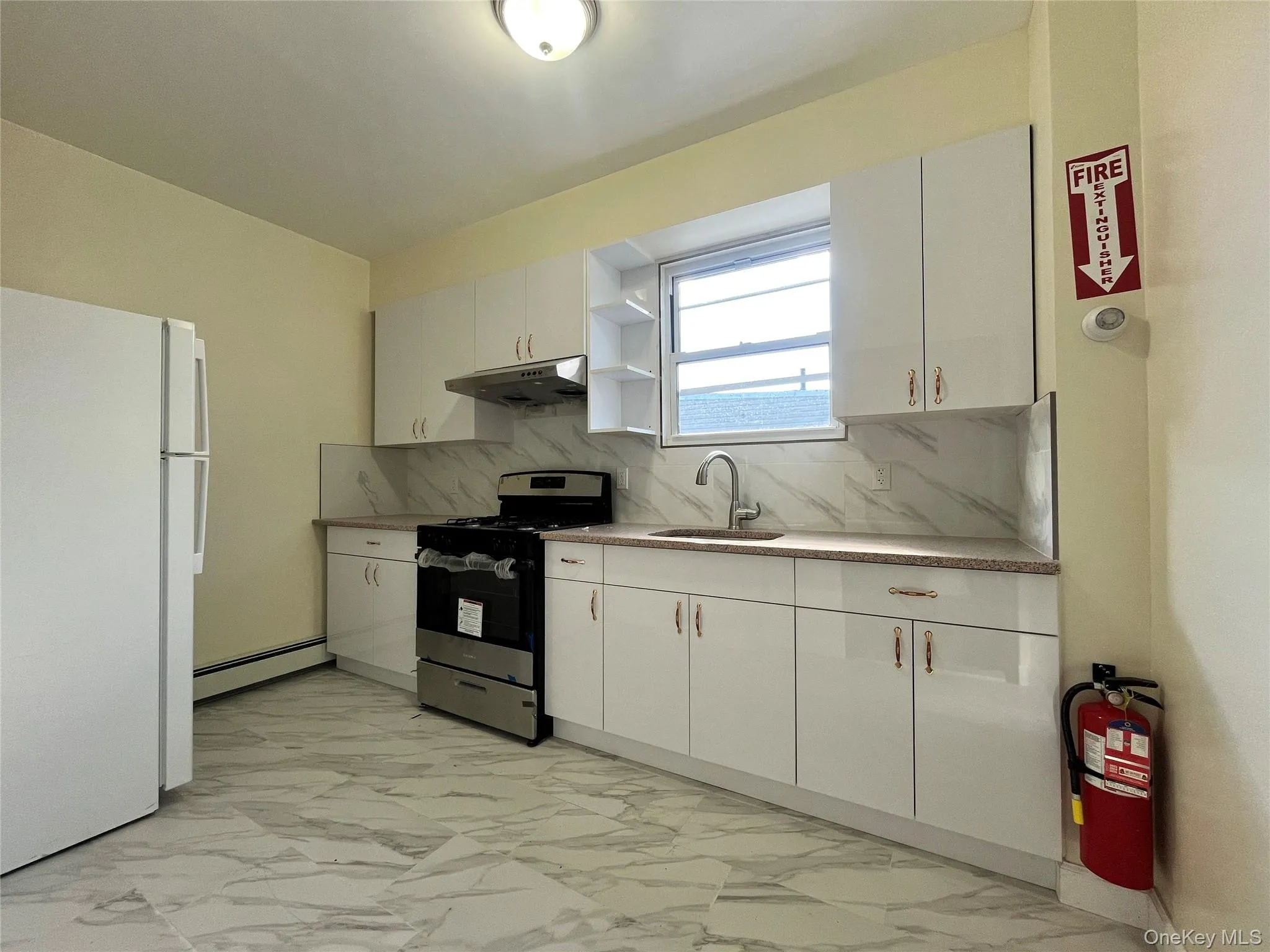 Kitchen with stainless steel gas stove, freestanding refrigerator, light marble finish flooring, white cabinetry, and backsplash Kitchen with stainless steel gas stove, freestanding refrigerator, light marble finish flooring, white cabinetry, and backsplash