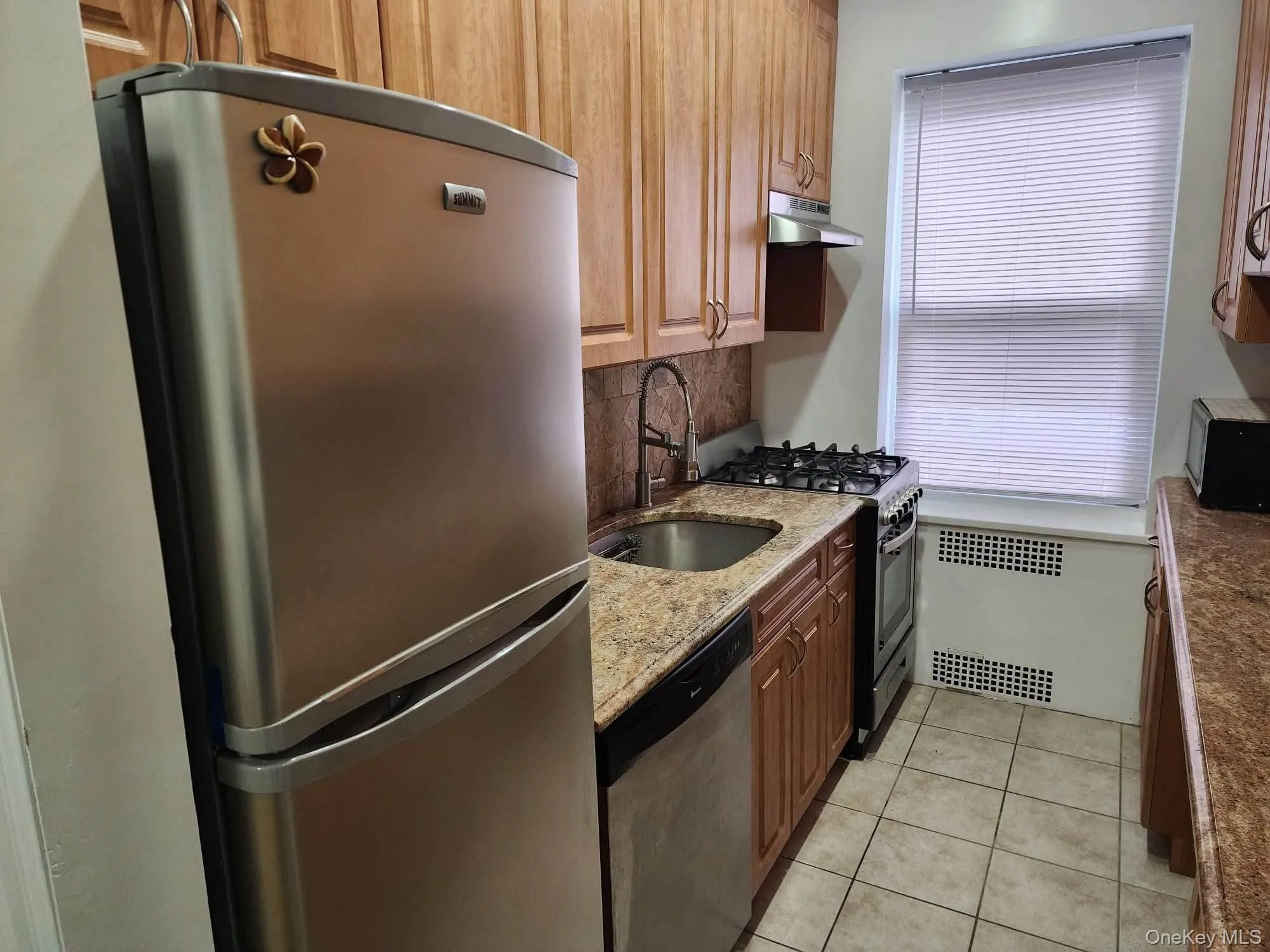 Kitchen featuring appliances with stainless steel finishes, light stone countertops, light tile patterned flooring, decorative backsplash, and under cabinet range hood Kitchen featuring appliances with stainless steel finishes, light stone countertops, light tile patterned flooring, decorative backsplash, and under cabinet range hood